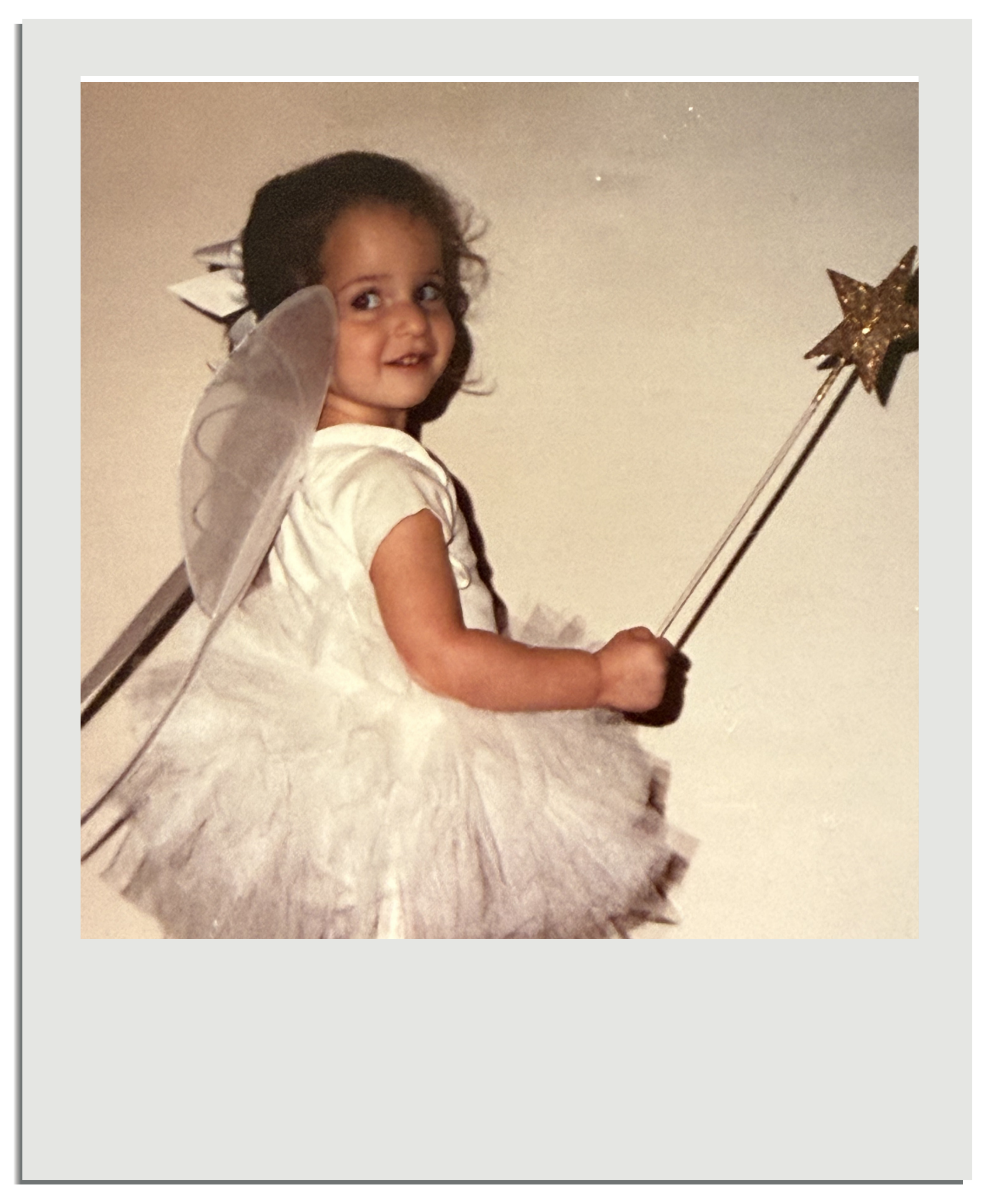 Young girl smiling and waving, wearing a fairy costume with wings and a star-shaped wand, standing on a fluffy rug in a neutral-colored room.