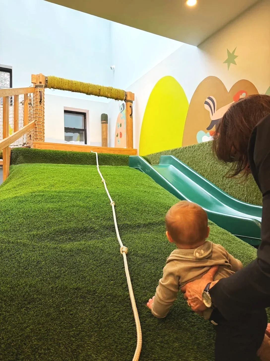 A young child on all fours at the bottom of an indoor play area slide, with an adult holding them from behind. The play area has a green artificial grass surface, a small slide, and colorful wall art.