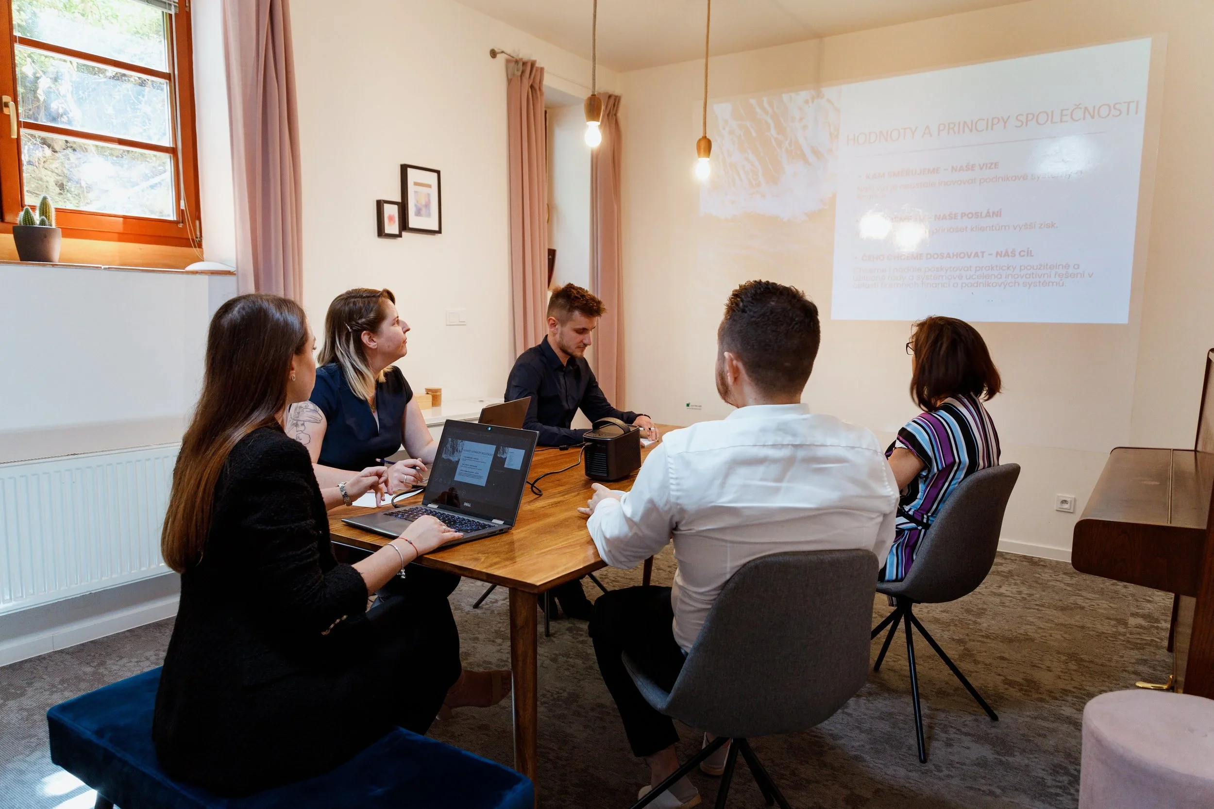 A group of five people attending a business meeting or presentation in a conference room, with a projector displaying a slide on the wall.