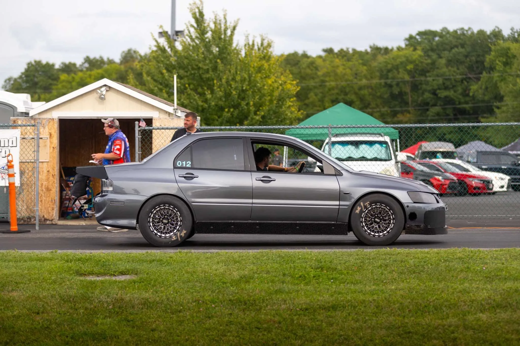 A gray sports car parked along a chain-link fence, with a woman inside and two men nearby, one walking past and the other standing behind the car.