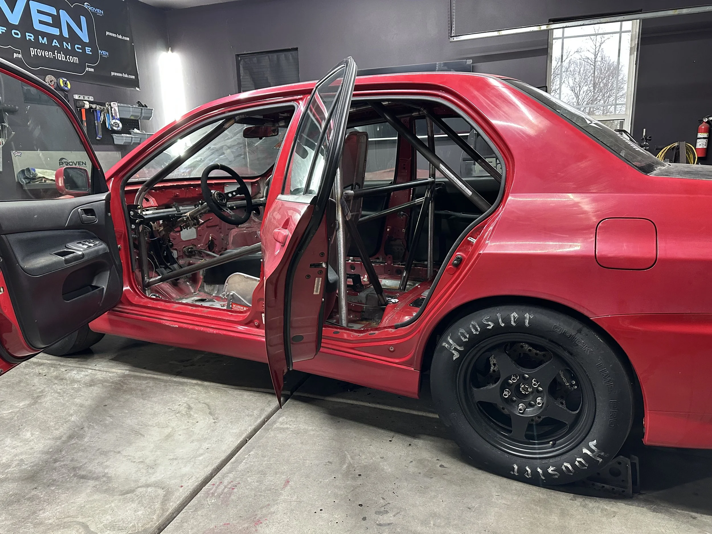 Red race car inside a garage, with doors open, showing the interior with a roll cage and racing seats. The garage has tools and equipment on the wall.