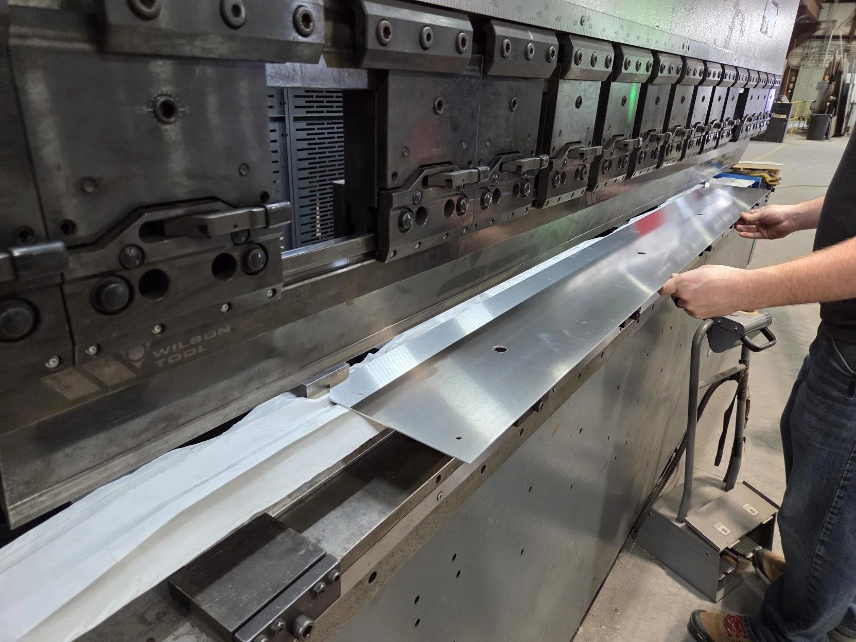 A person working on a large industrial metal press machine in a factory, holding a sheet of metal.