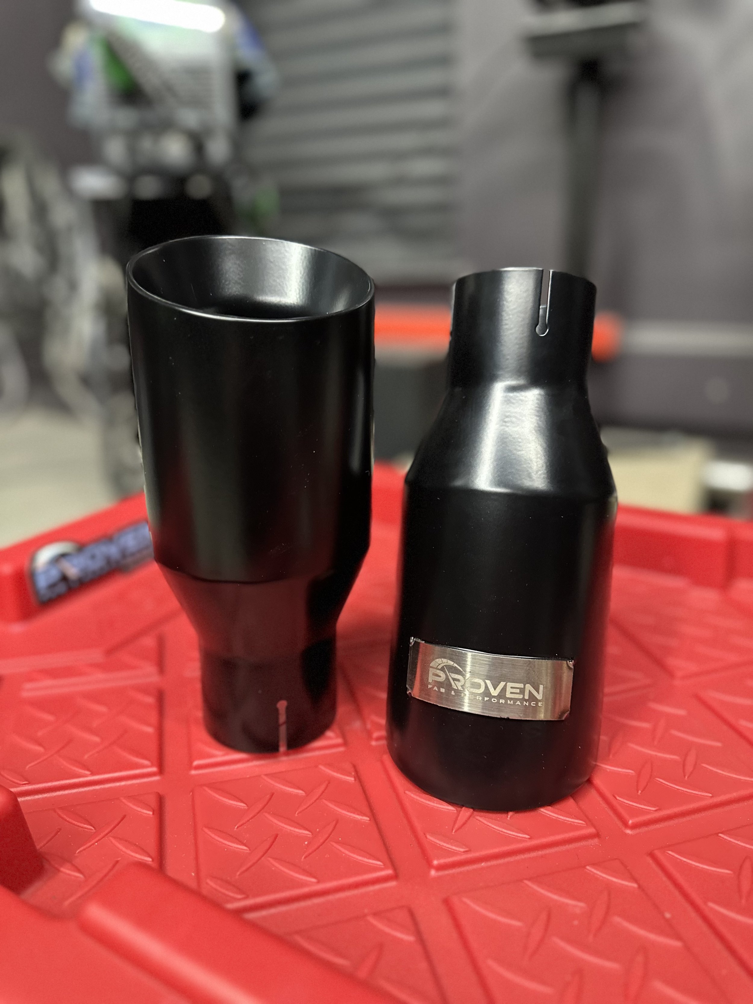 Close-up of two black motorcycle shock absorbers on a red diamond-patterned surface, one marked with a Proven logo.