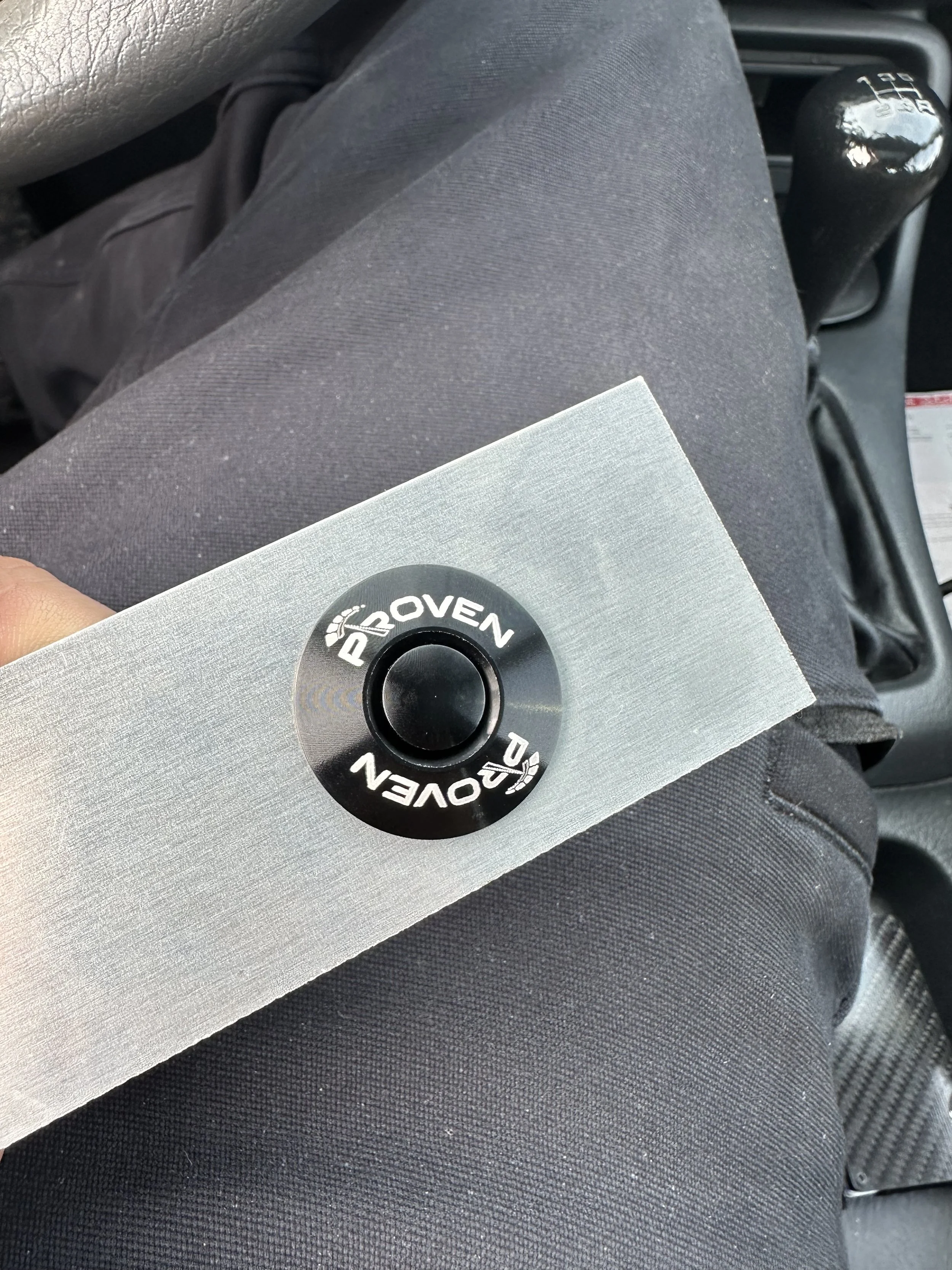 Close-up of a manual gear shift in a car, with a person holding a metal circle with the word 'PROVEN' on it, sitting on black fabric seat.