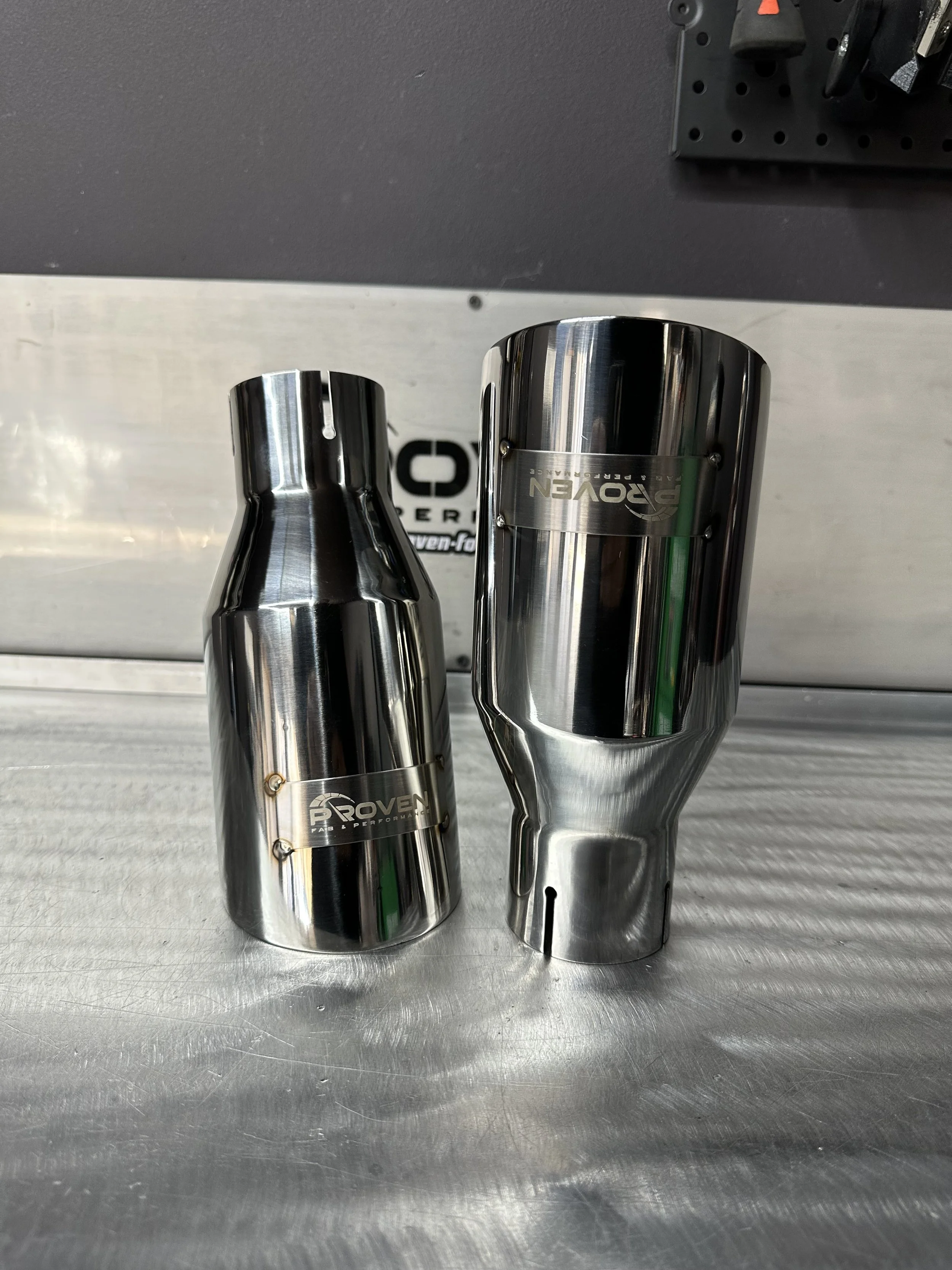 Two shiny metallic motorcycle exhaust pipes placed on a metal surface with a black wall and a black pegboard holding tools in the background.