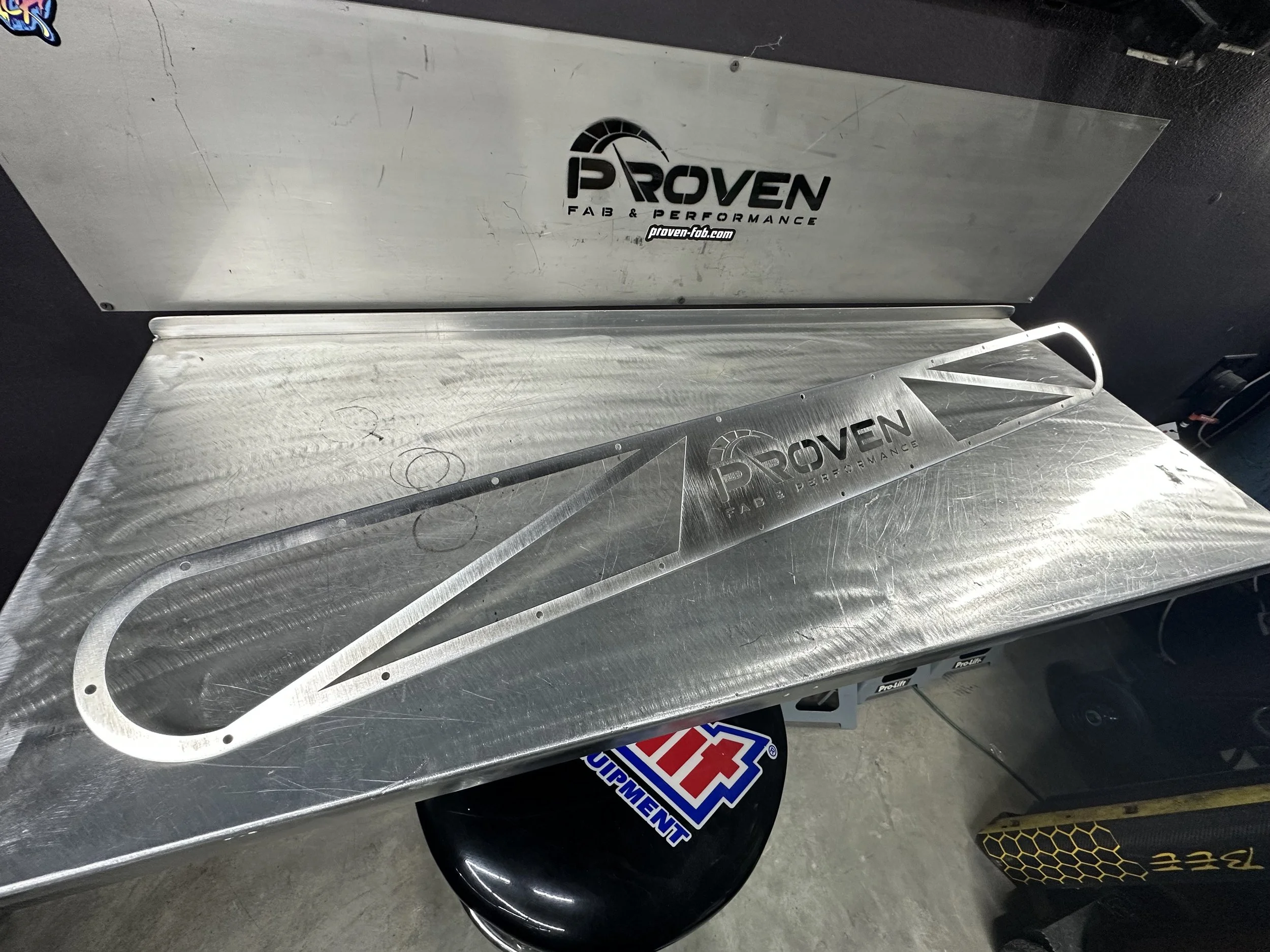 Metal workshop table with Proven Fab & Performance logo, metal car part template, and black helmet with stickers.
