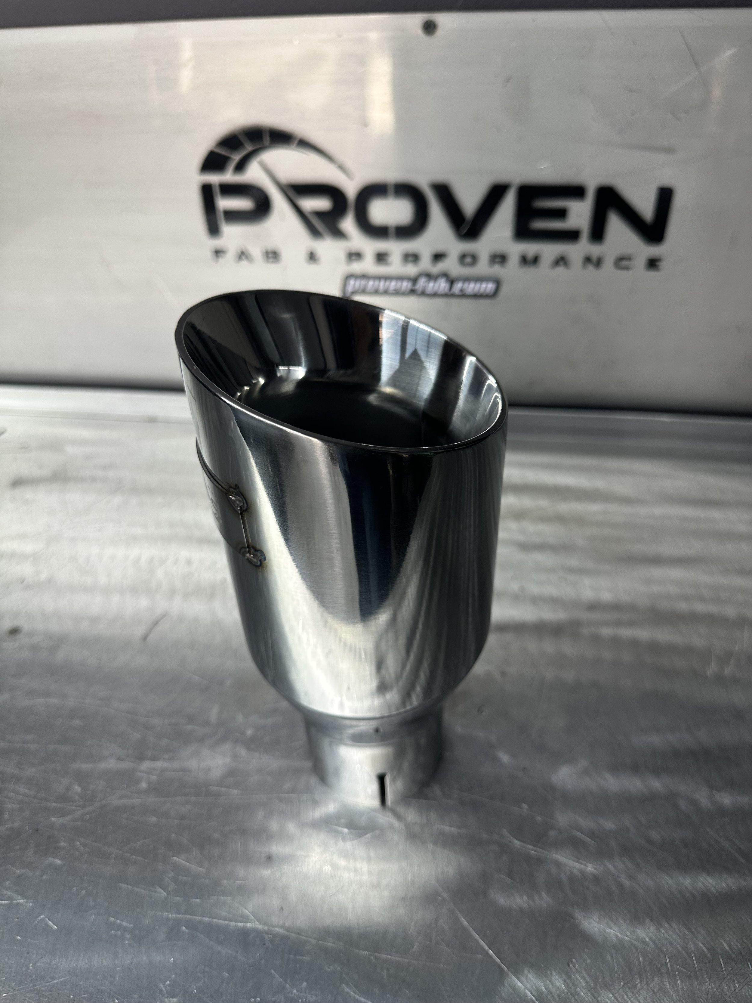 Metal exhaust pipe with a shiny, reflective surface in front of a metal surface with a "Proven Fab & Performance" logo etched on it.