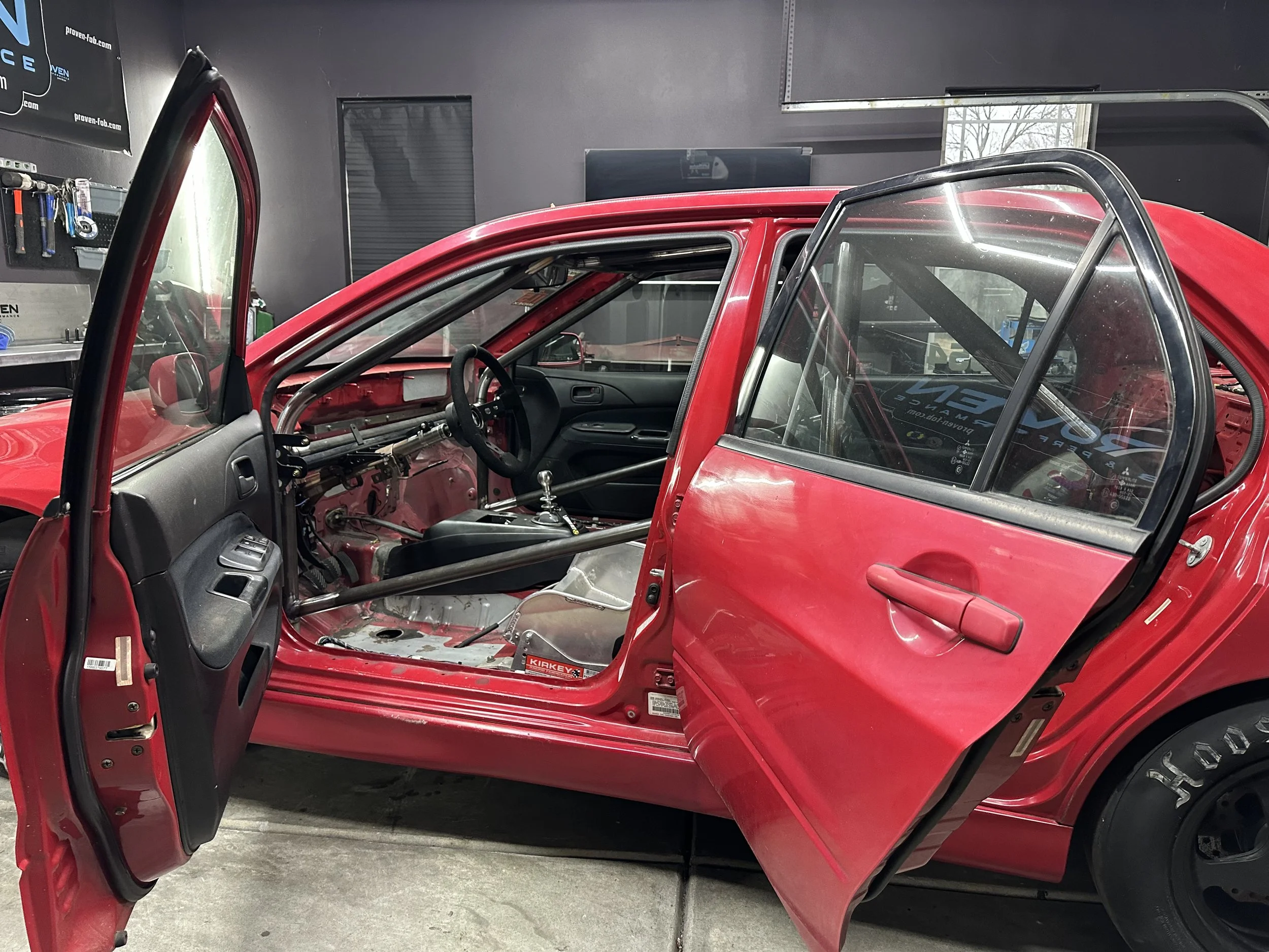 A red race car with no interior, exposing the roll cage and other racing modifications inside, parked in a garage.