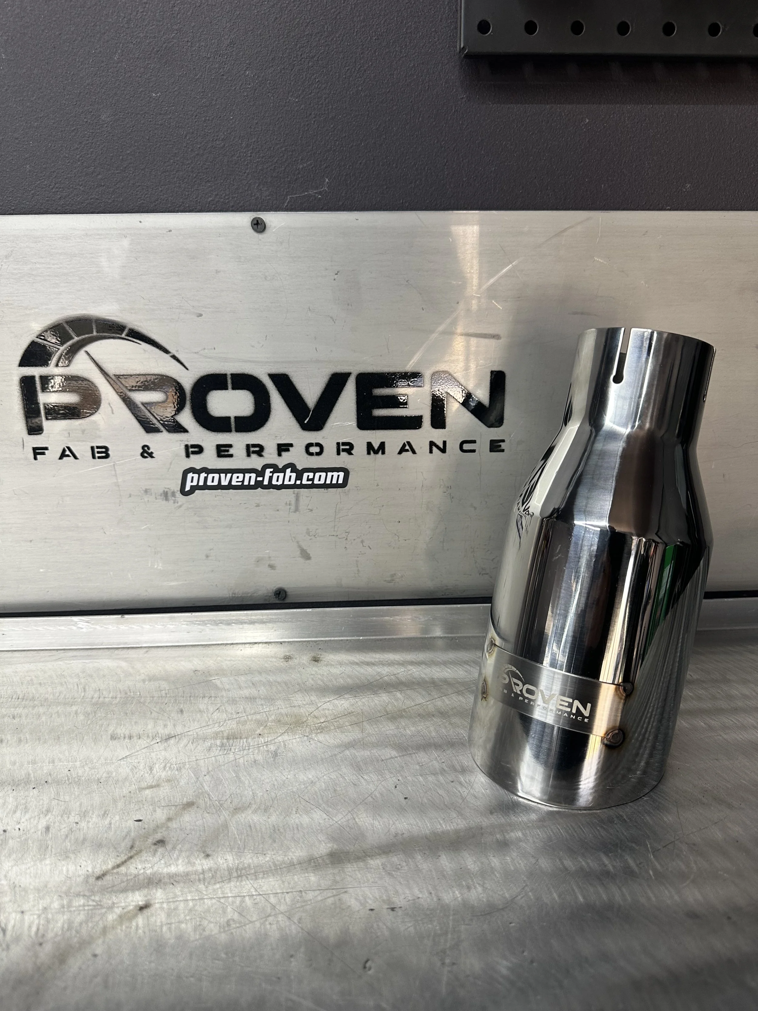 Close-up of a metallic box and a shiny, cylindrical component with 'PROVEN' logo, placed on a metal surface, with a black textured background.