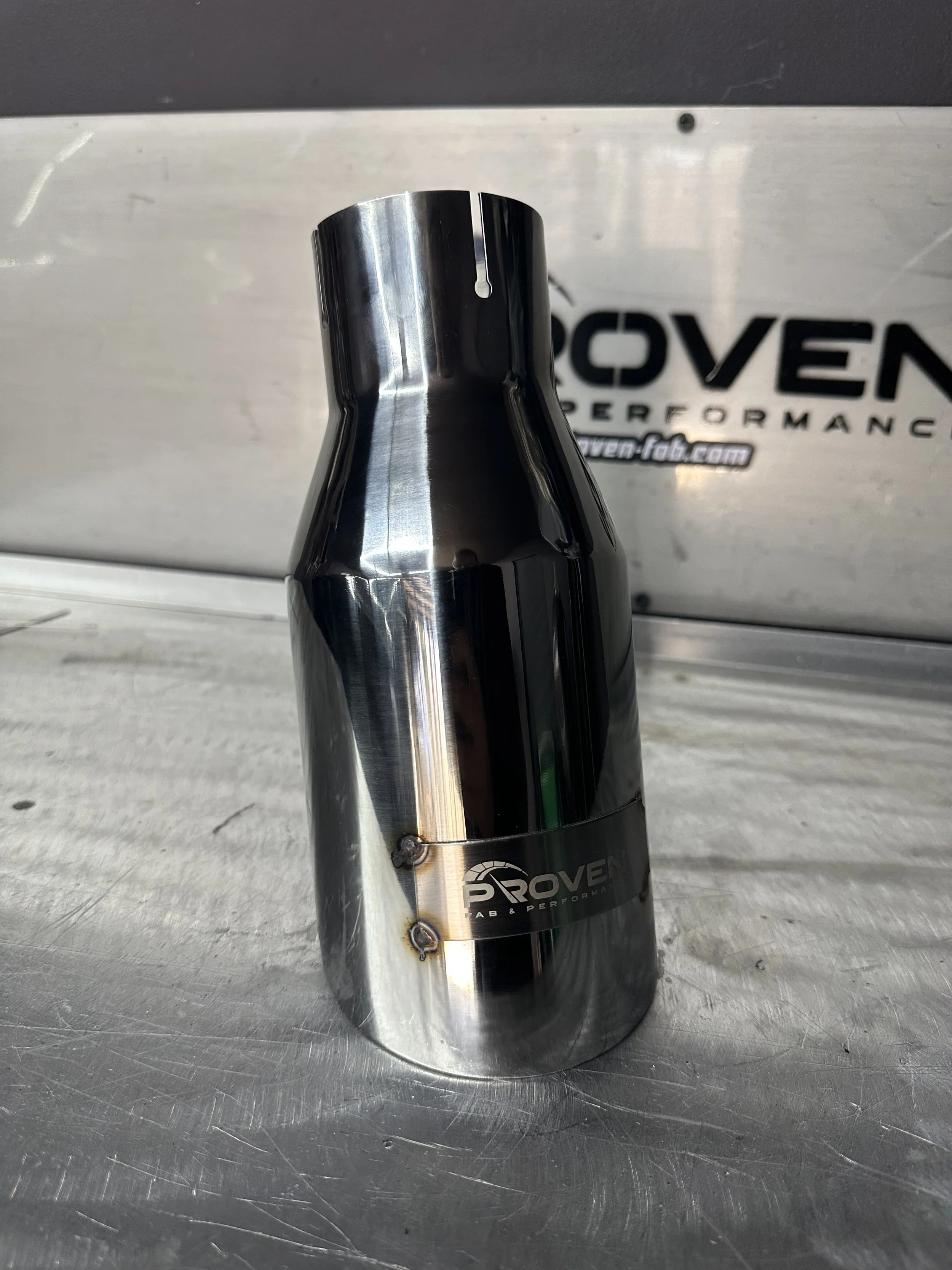 Close-up of a polished metal exhaust pipe with a 'Proven' logo, resting on a metal surface with a background showing a Proven Performance sign.