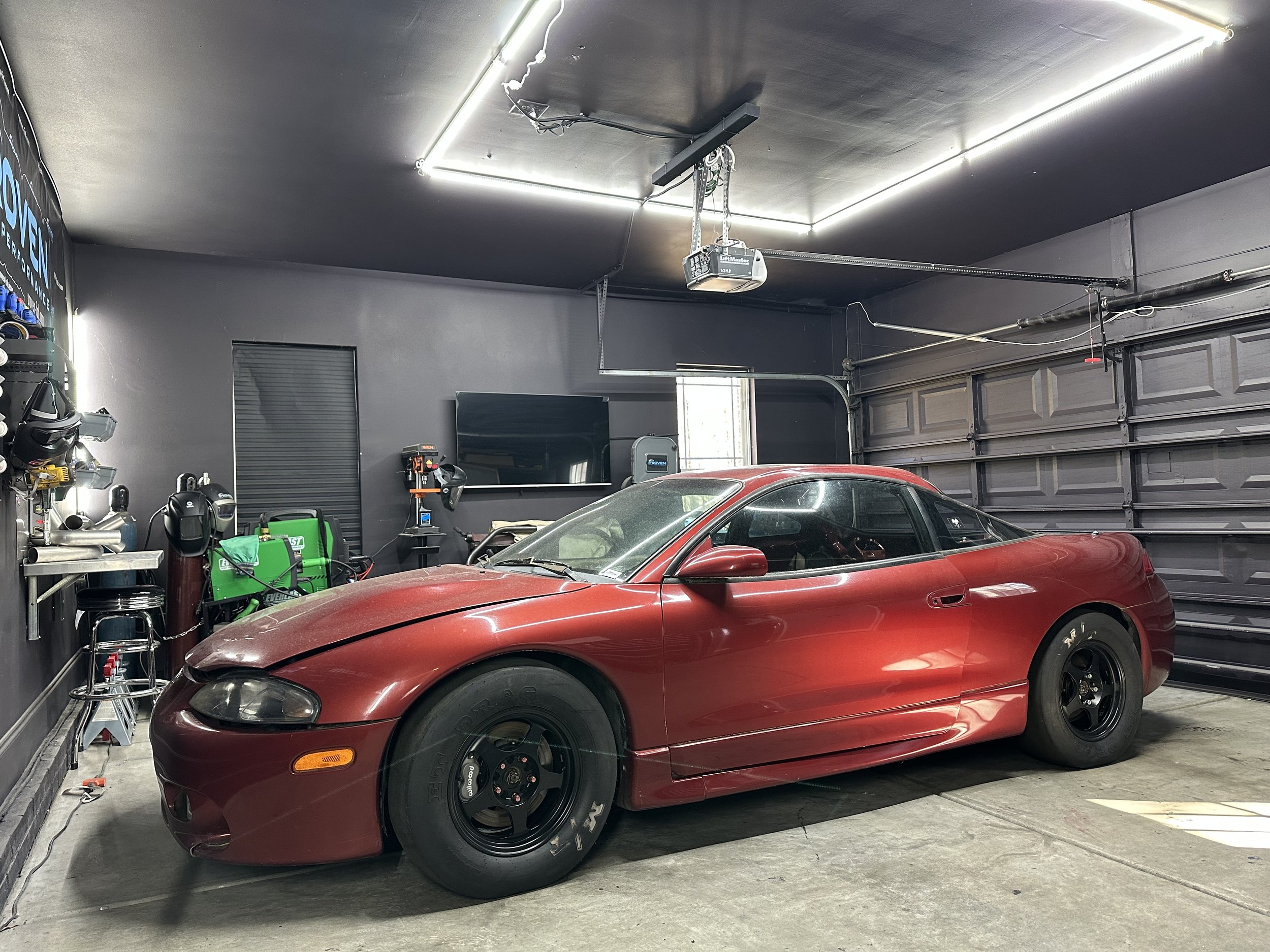 A red sports car is parked inside a garage with gray walls and a gray ceiling. The garage contains tools and equipment on shelves and a television mounted on the wall. There is a garage door in the background and a ceiling-mounted garage door opener.