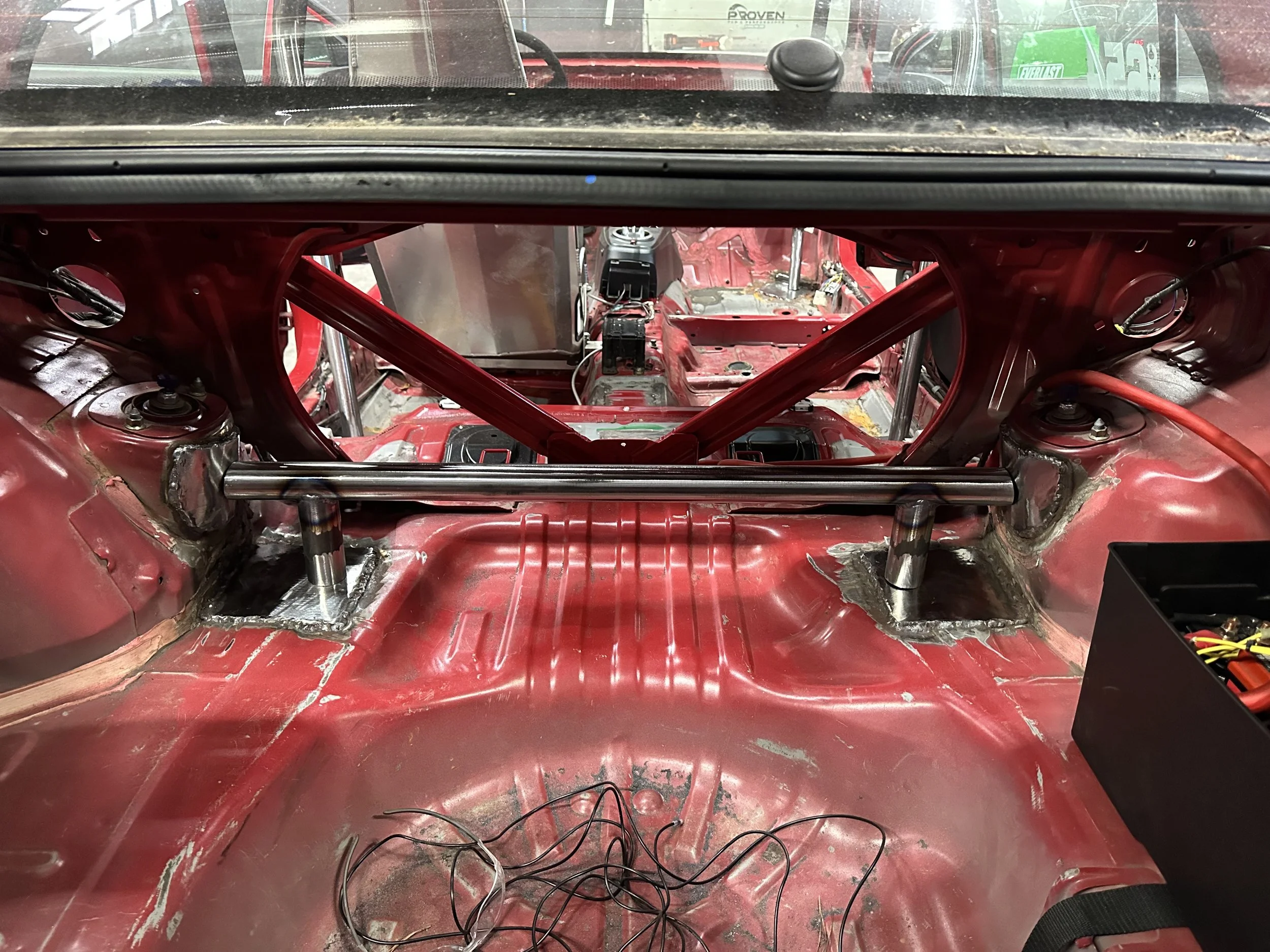 View from inside a red race car's empty interior with no seats, showing the bare metal and wiring, looking towards the dashboard area and through the windshield to other vehicles outside.