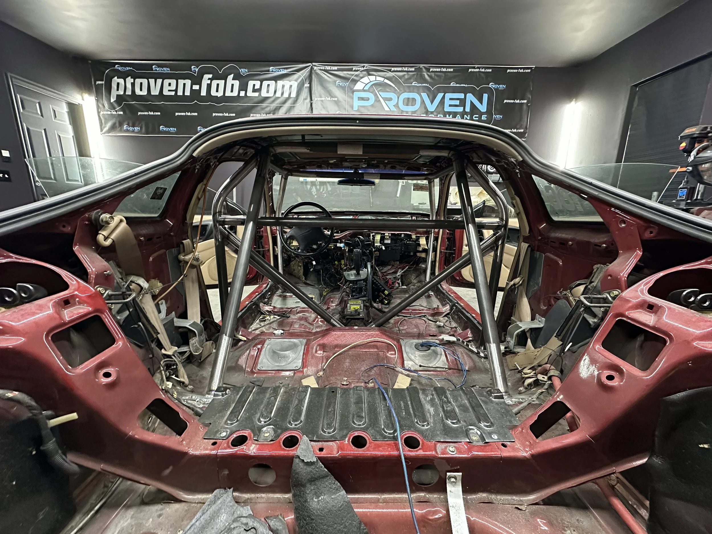 The interior of a race car chassis under construction, featuring a metal roll cage and exposed wiring, with a banner in the background displaying the website 'proven-fab.com'.