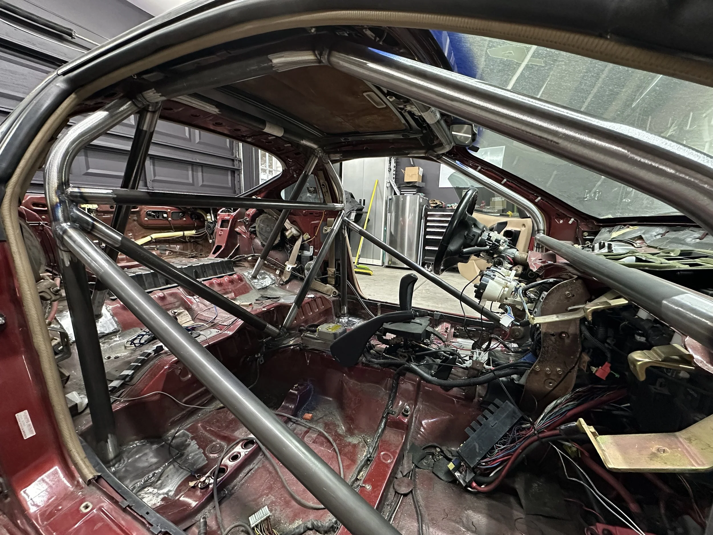 The interior of a car with its roof and interior panels removed, showing a roll cage, wiring, and metal framework in a garage or workshop.