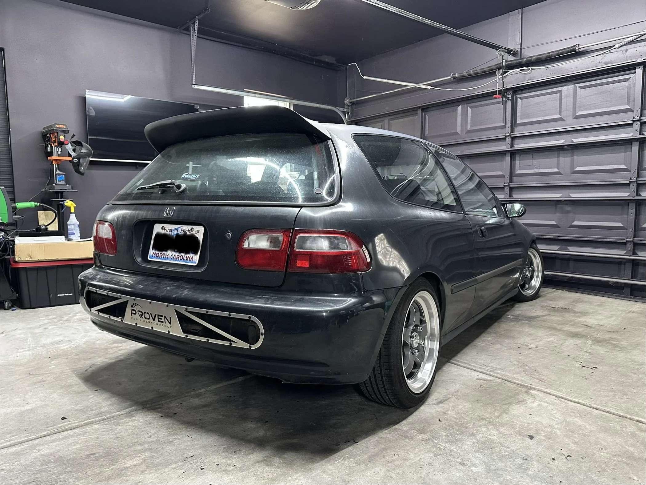 A black Honda Civic hatchback parked inside a garage with a raised door. The car has aftermarket wheels, a custom front bumper, and a spoiler. The garage has a concrete floor, gray walls, and various tools and supplies visible on the left side.