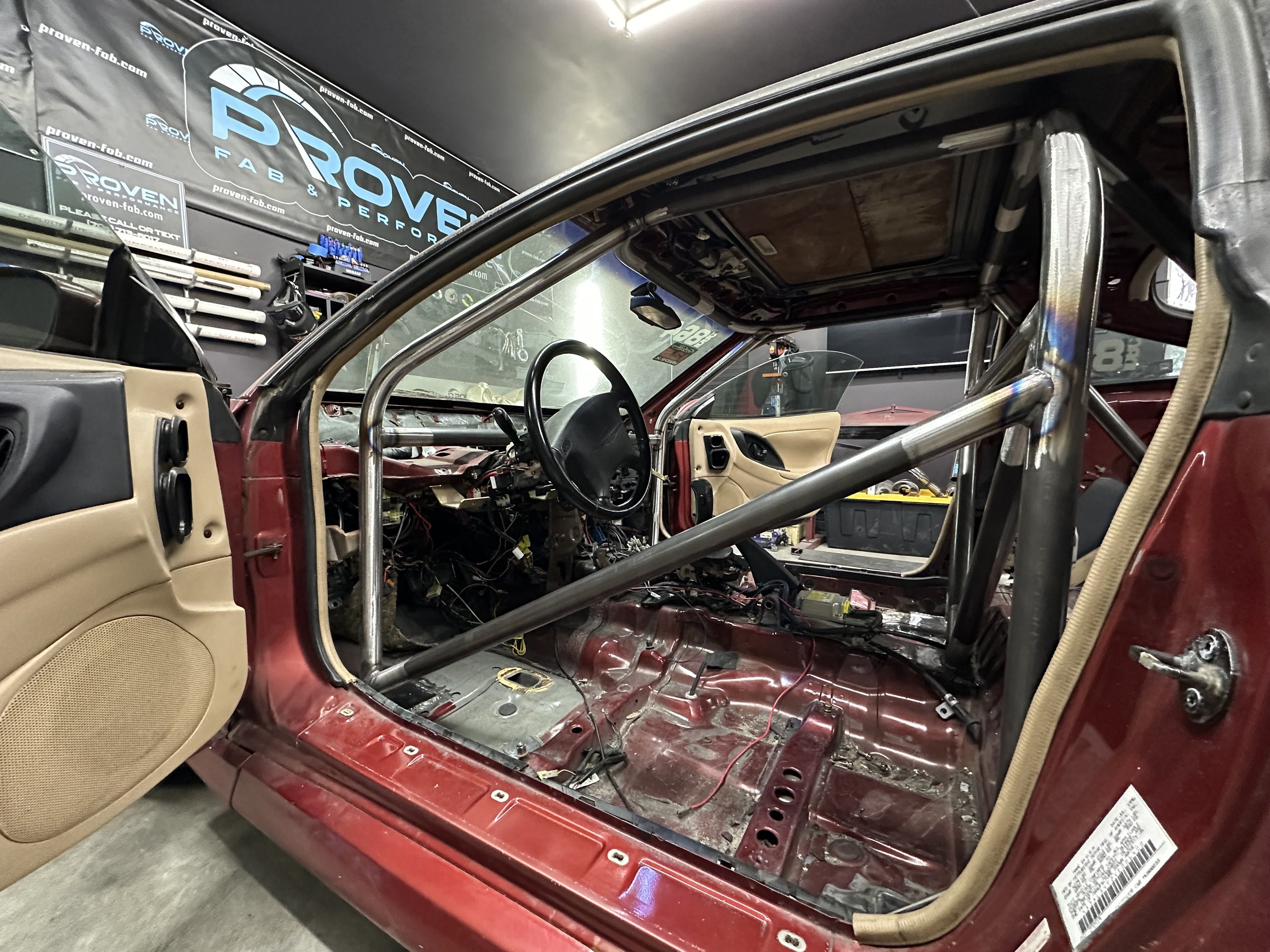Interior of a race car in the process of being assembled or repaired, with exposed metal framework, wiring, and partial dashboard, inside a workshop with a banner reading 'Proven-Fab' in the background.