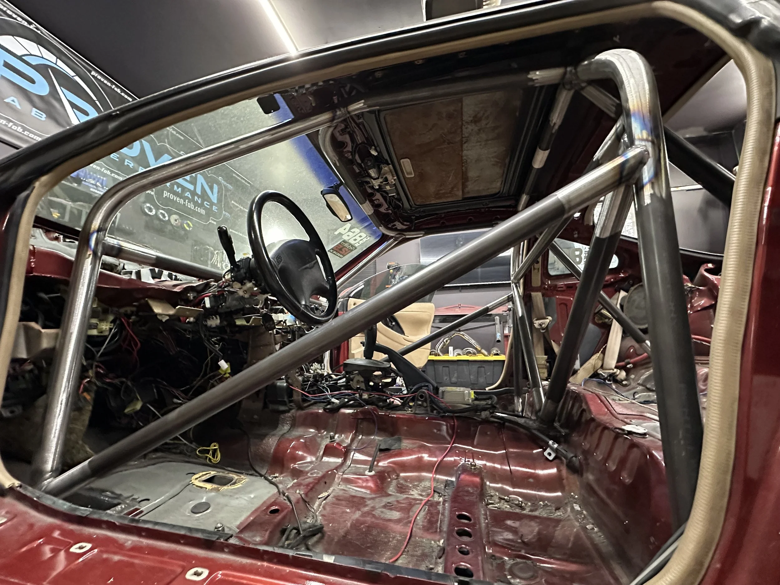 Interior of a race car with exposed metal framework, wiring, and no interior panels, showing the dashboard, steering wheel, and roll cage.