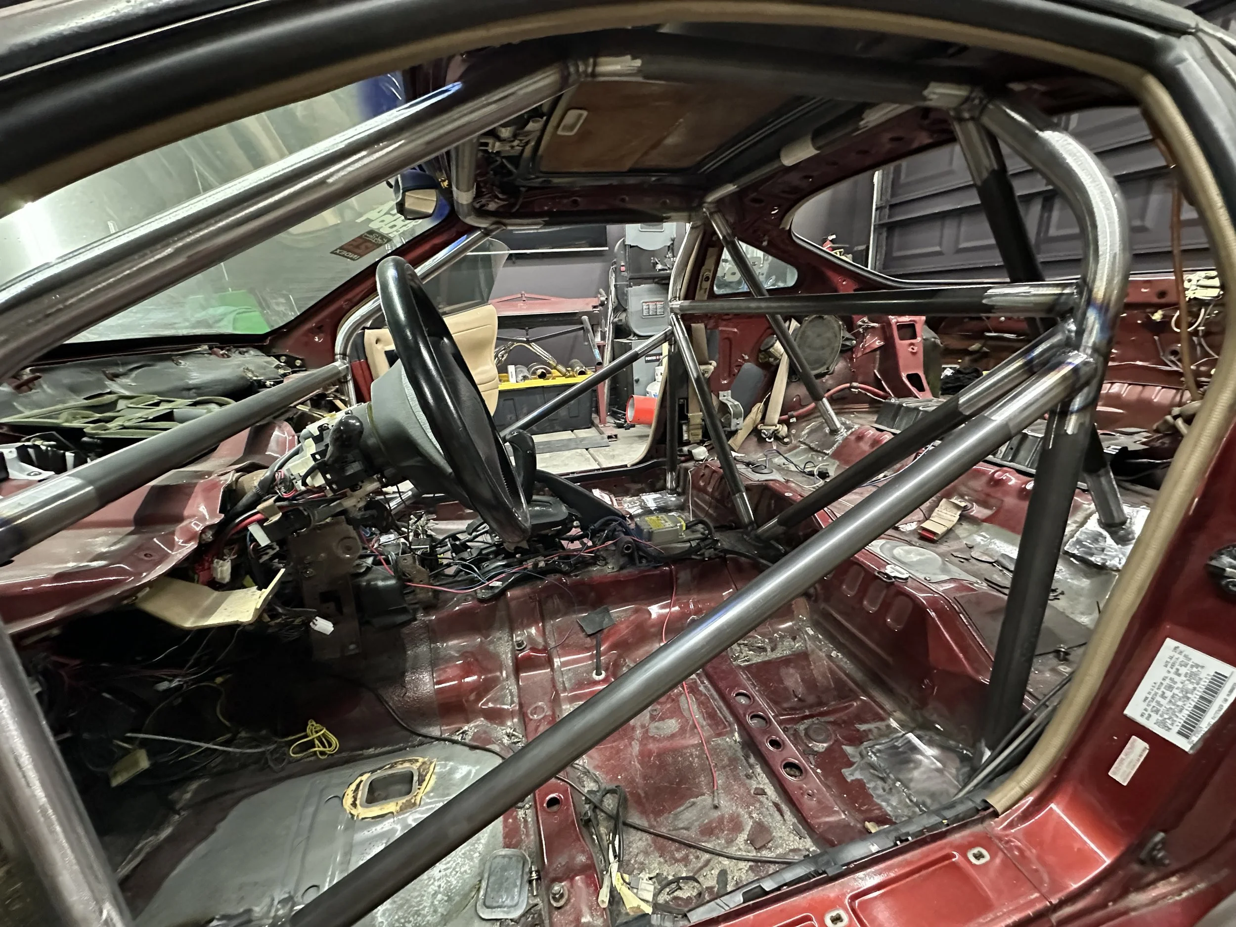 The interior of a race car under construction, showing a roll cage and bare metal surfaces, with various wires and components visible.