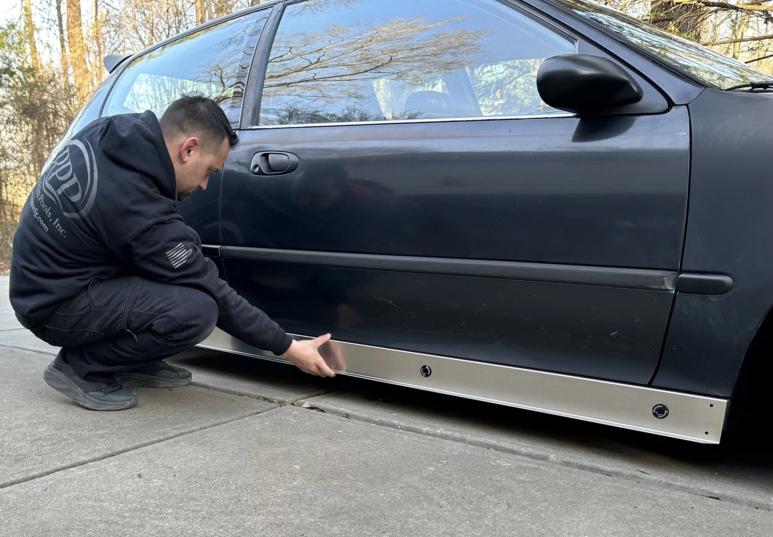 A man crouching down next to a black car, working on or inspecting the undercarriage, in a driveway with trees in the background.
