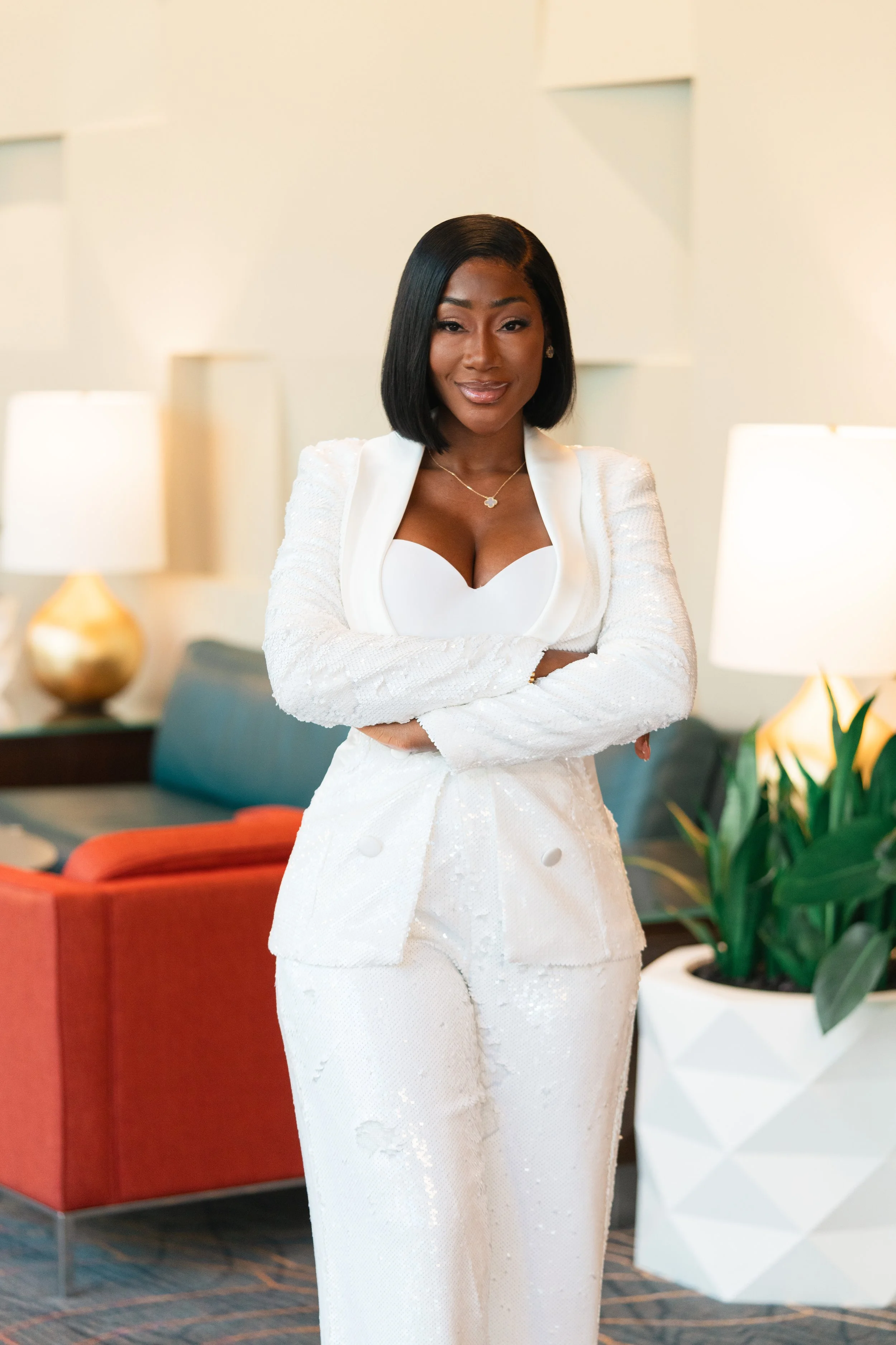 A confident woman standing with crossed arms, wearing a white suit and top, in a modern, well-lit indoor setting with lamps, a plant, and contemporary furniture.