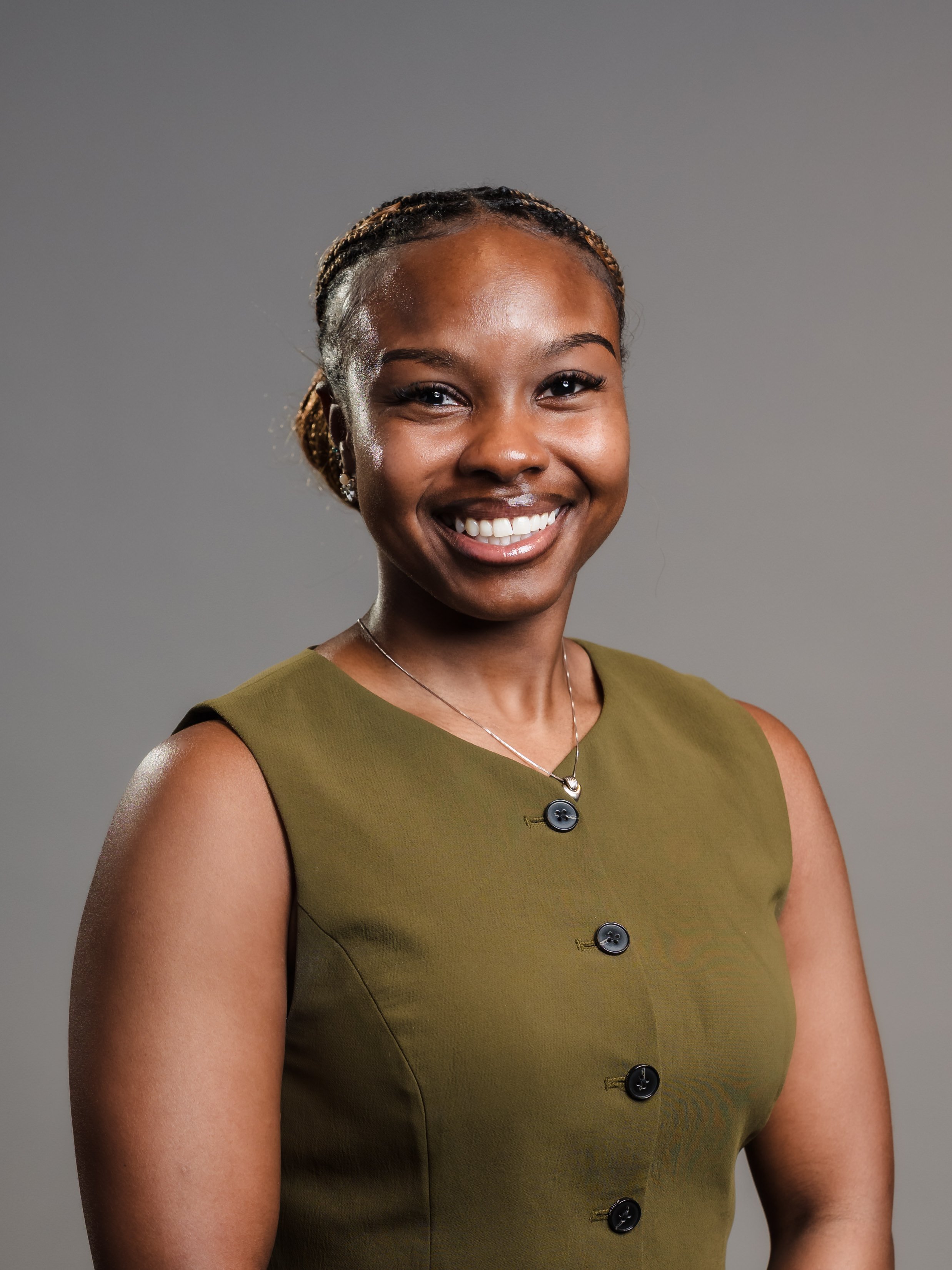 A woman with dark skin and braided hair smiling, wearing a sleeveless olive green dress and a necklace, against a plain gray background.