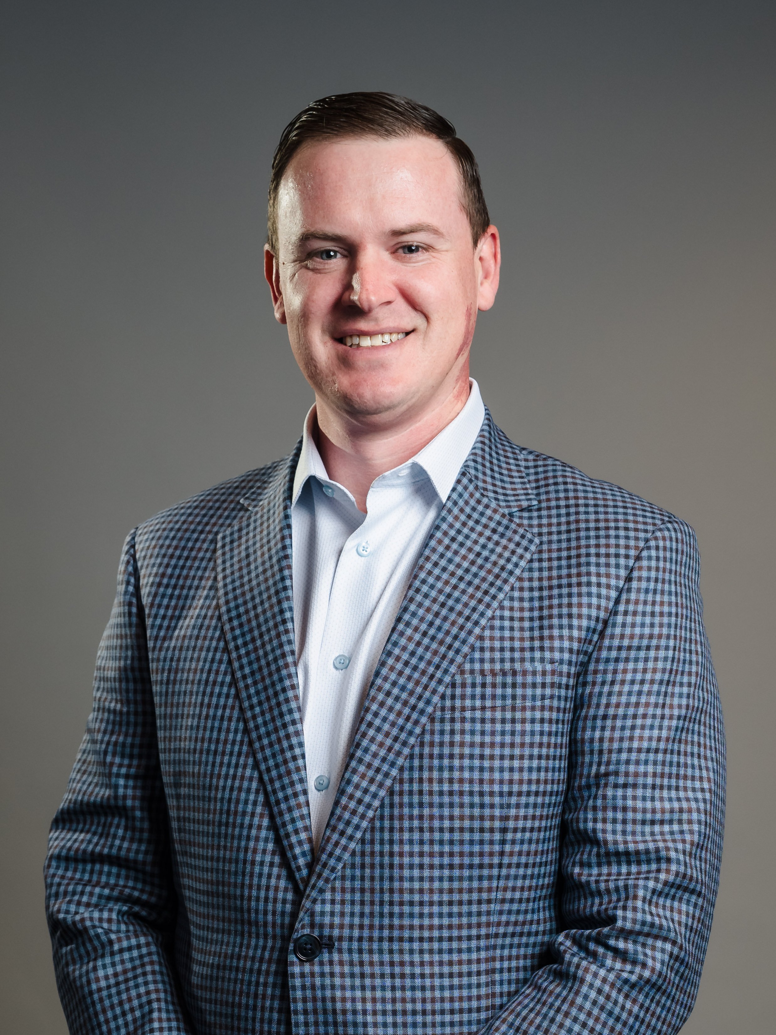 A young man with short dark hair, wearing a checkered blazer over a white dress shirt, smiling at the camera against a plain gray background.