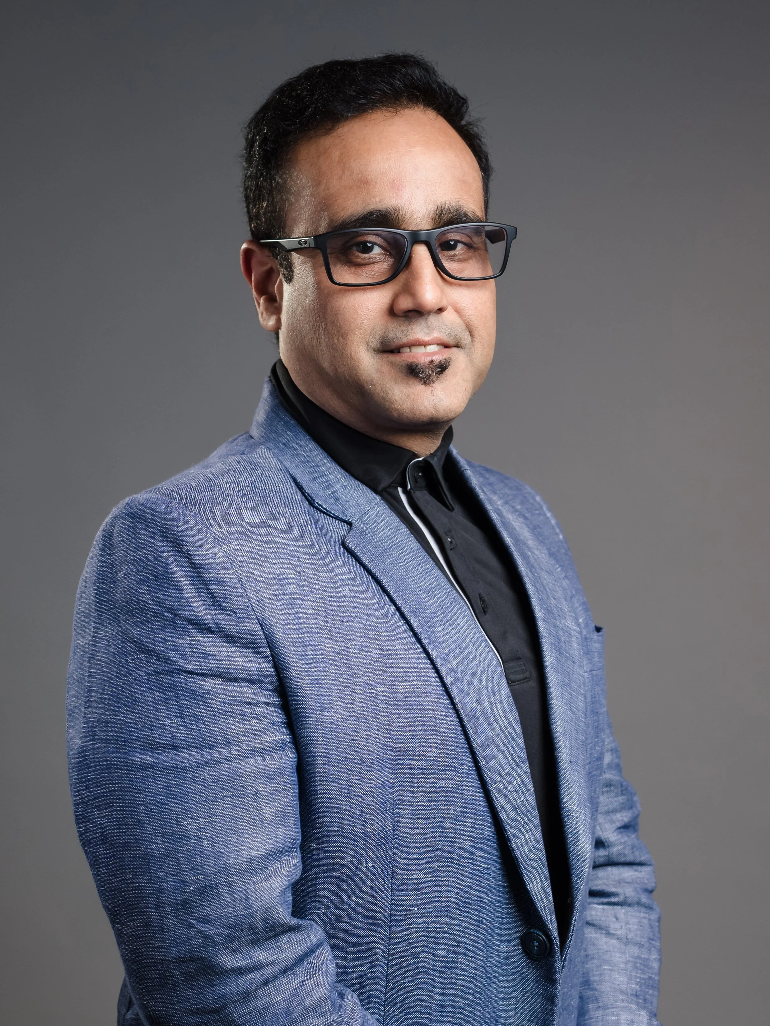 A man in a blue blazer, black shirt, and glasses posing for a professional portrait against a gray background.