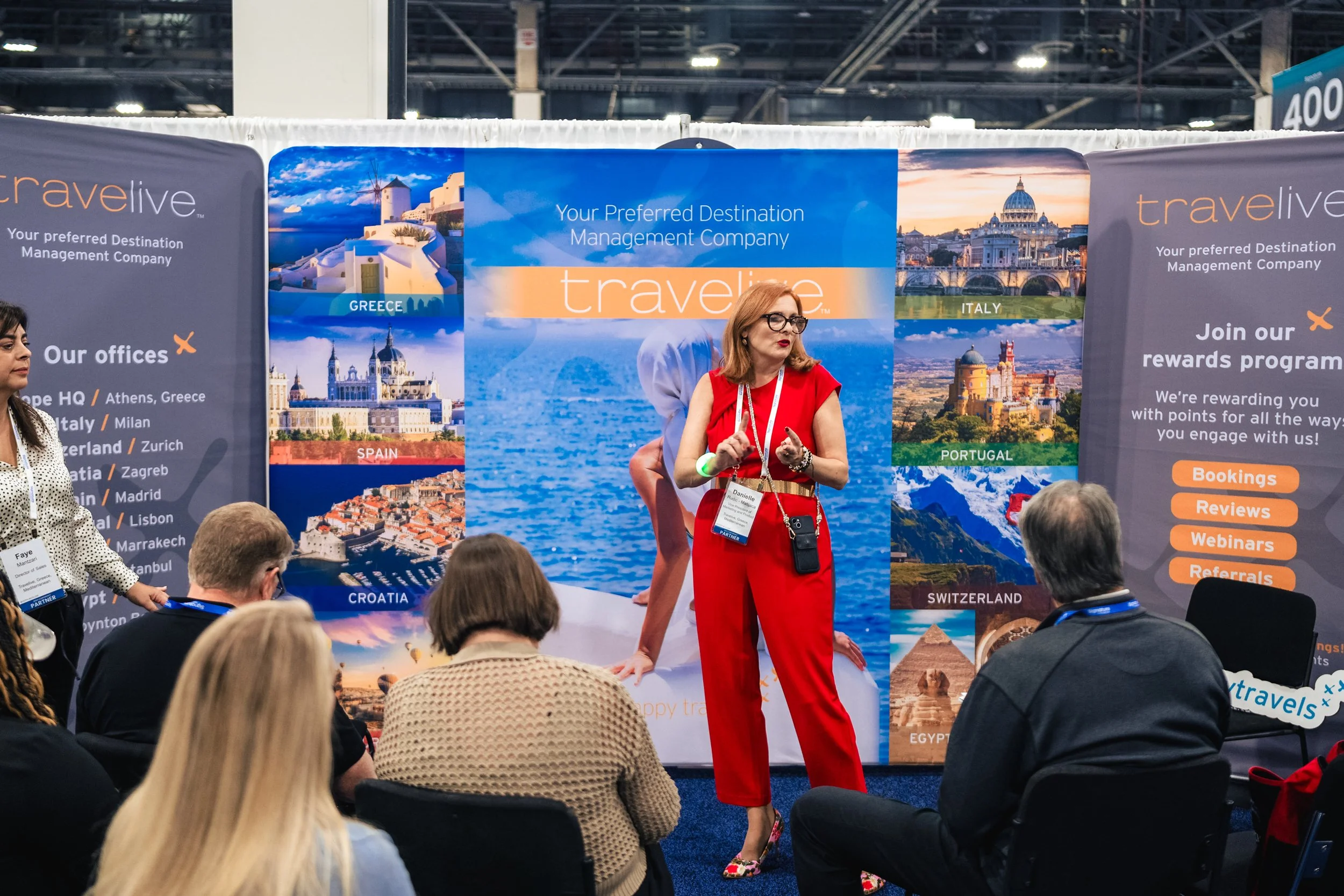 A woman in red pants and a red sleeveless top is giving a presentation at a travel expo, standing in front of a large booth with banners showcasing various European countries and travel destinations. She is holding a microphone and has a badge around