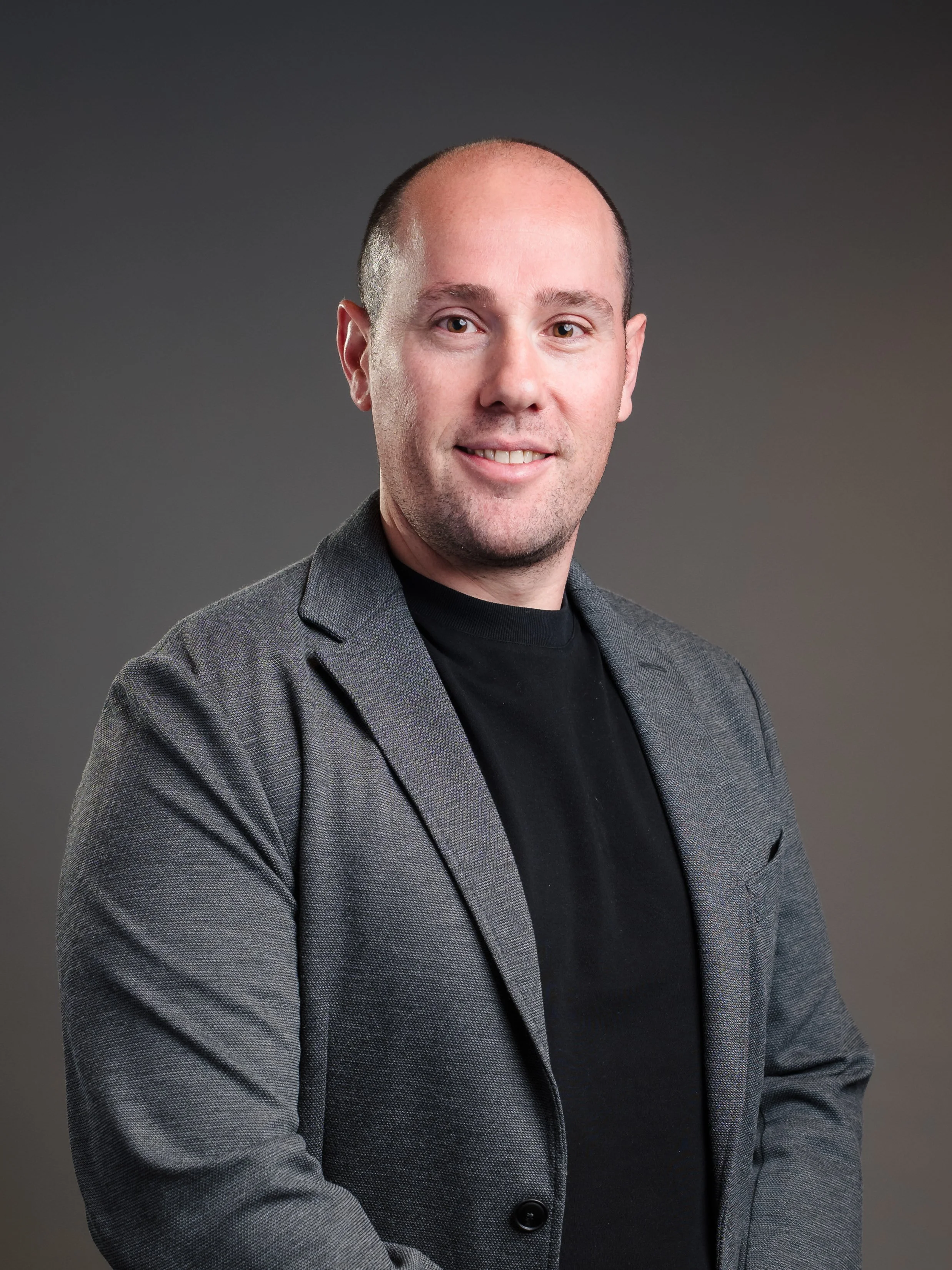 A professional portrait of a man with a confident smile, wearing a gray blazer and black shirt, with a neutral gray background.