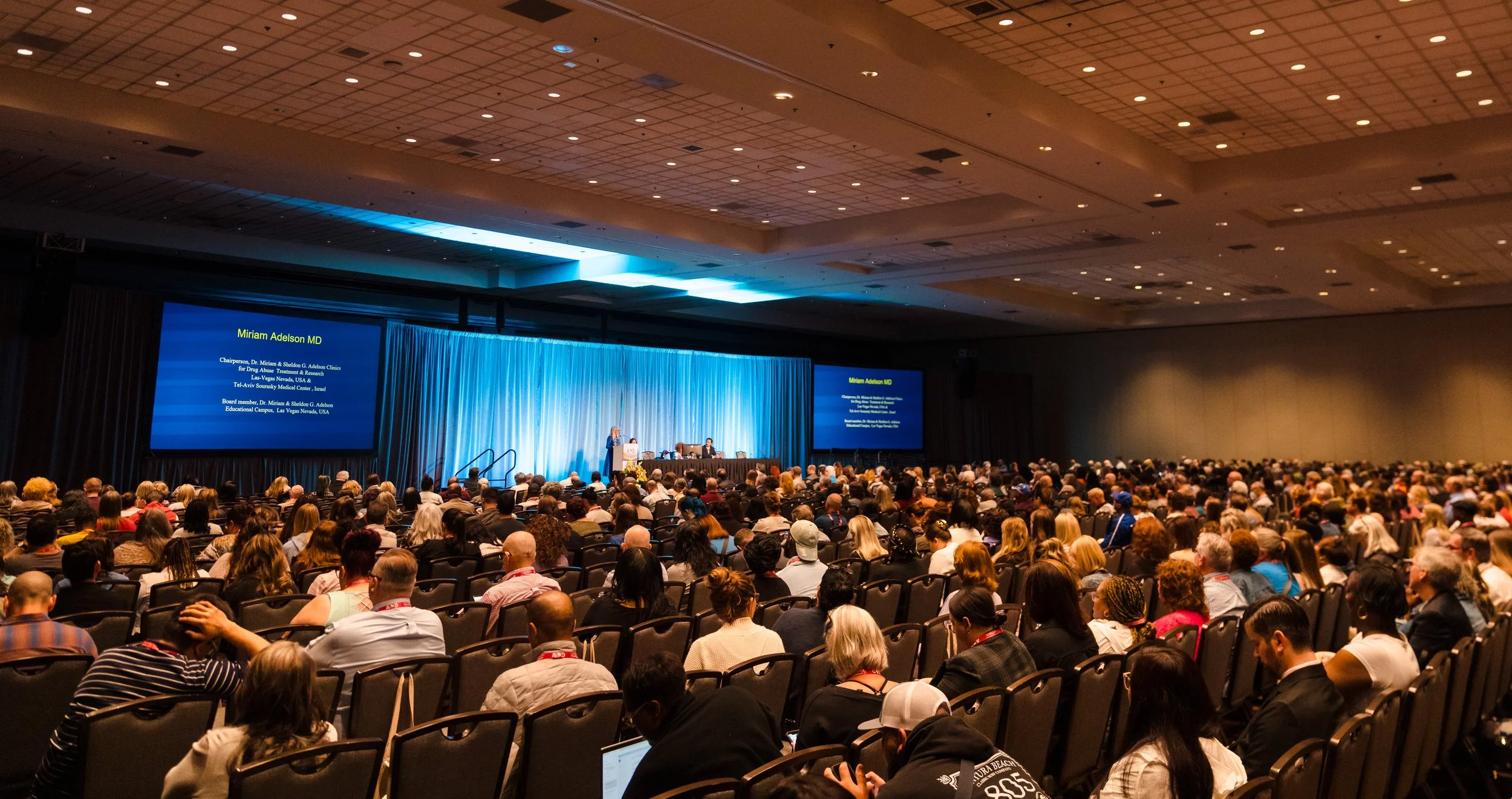 A large conference room filled with a diverse audience listening to a speaker on stage. The stage has a blue curtain backdrop and two large screens displaying the speaker's name, Miriam Adelson MD, and details about her credentials.