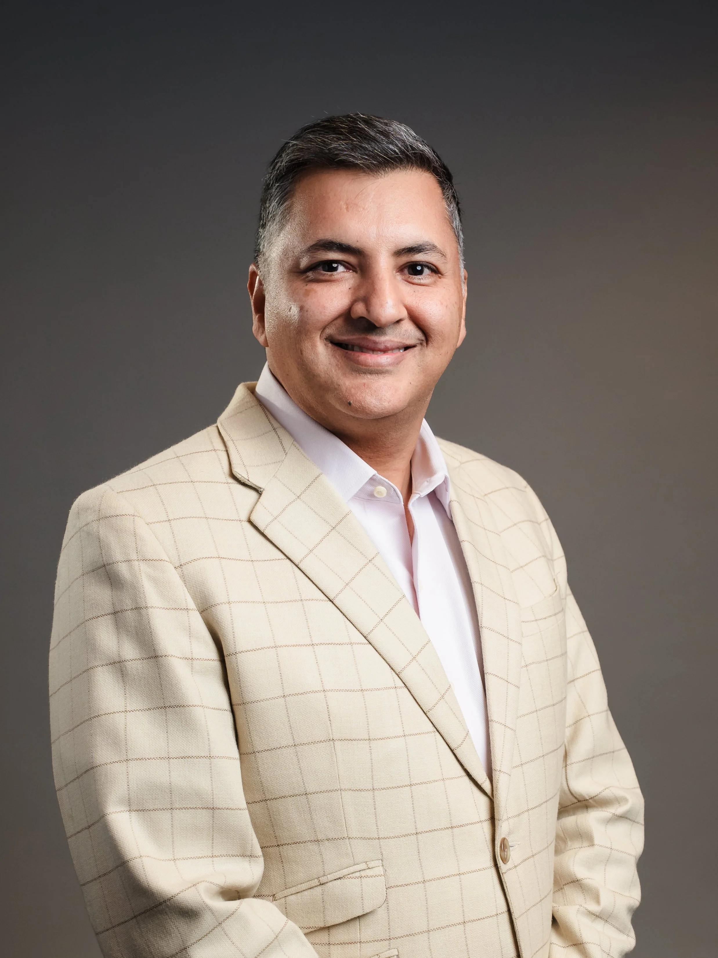 A portrait of a man in a beige checkered blazer and a white shirt against a gradient gray background.