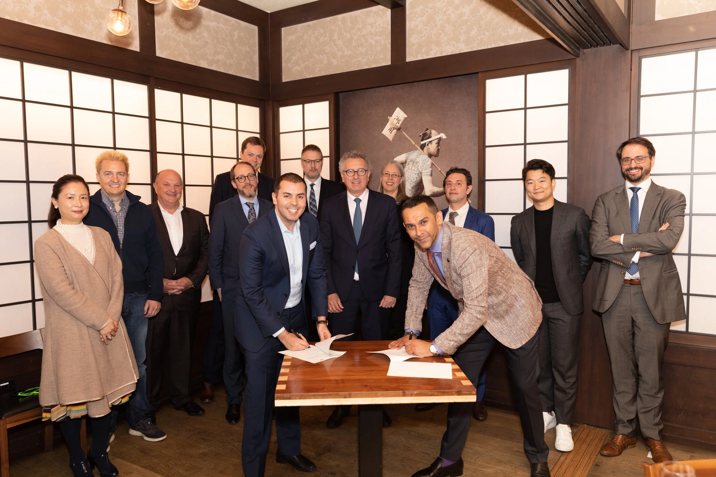 A group of professionals, including men and women, gathered in a room with traditional Japanese sliding doors, some standing behind and some leaning over a table with documents, engaging in a signing or agreement. The group appears happy and confiden