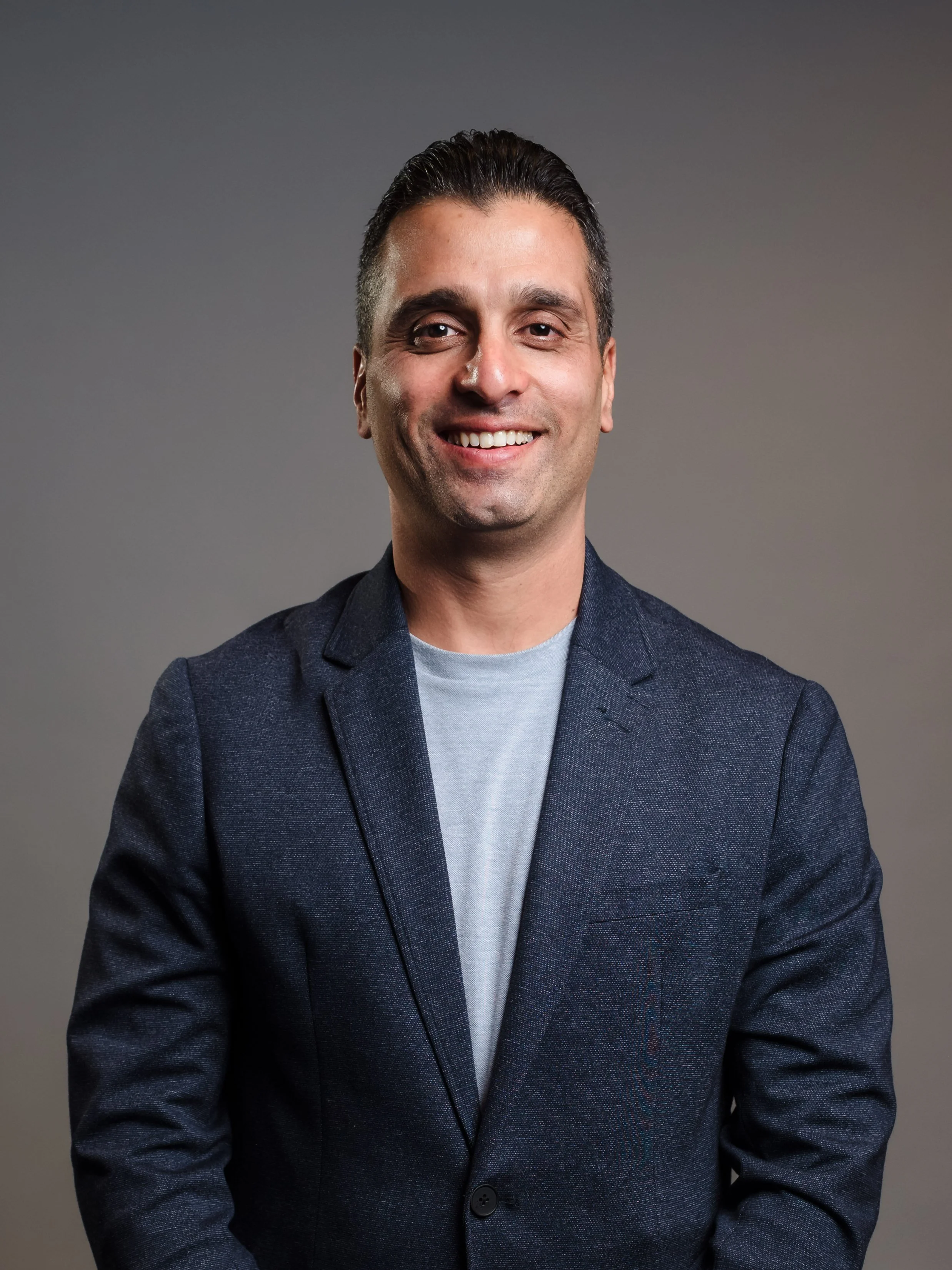 A smiling man with dark hair and a light complexion wearing a dark blazer and a light gray shirt, standing against a plain gray background.