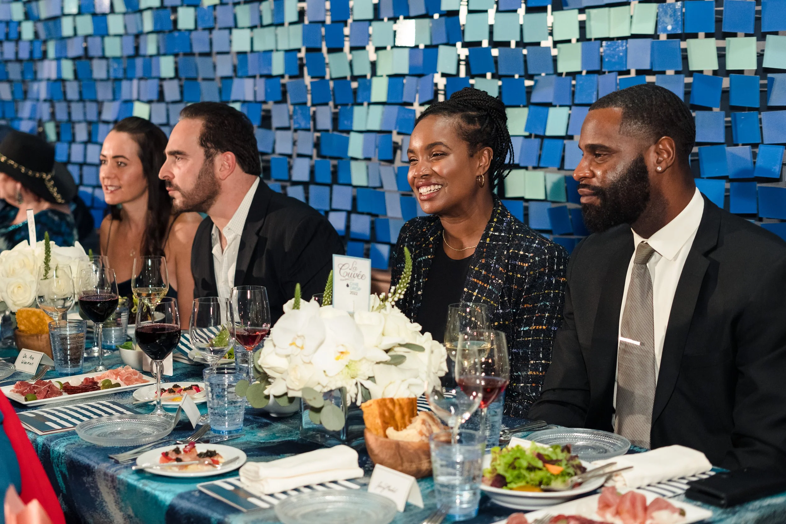 People sitting at a decorated dinner table during a formal event, with a blue geometric patterned wall in the background.