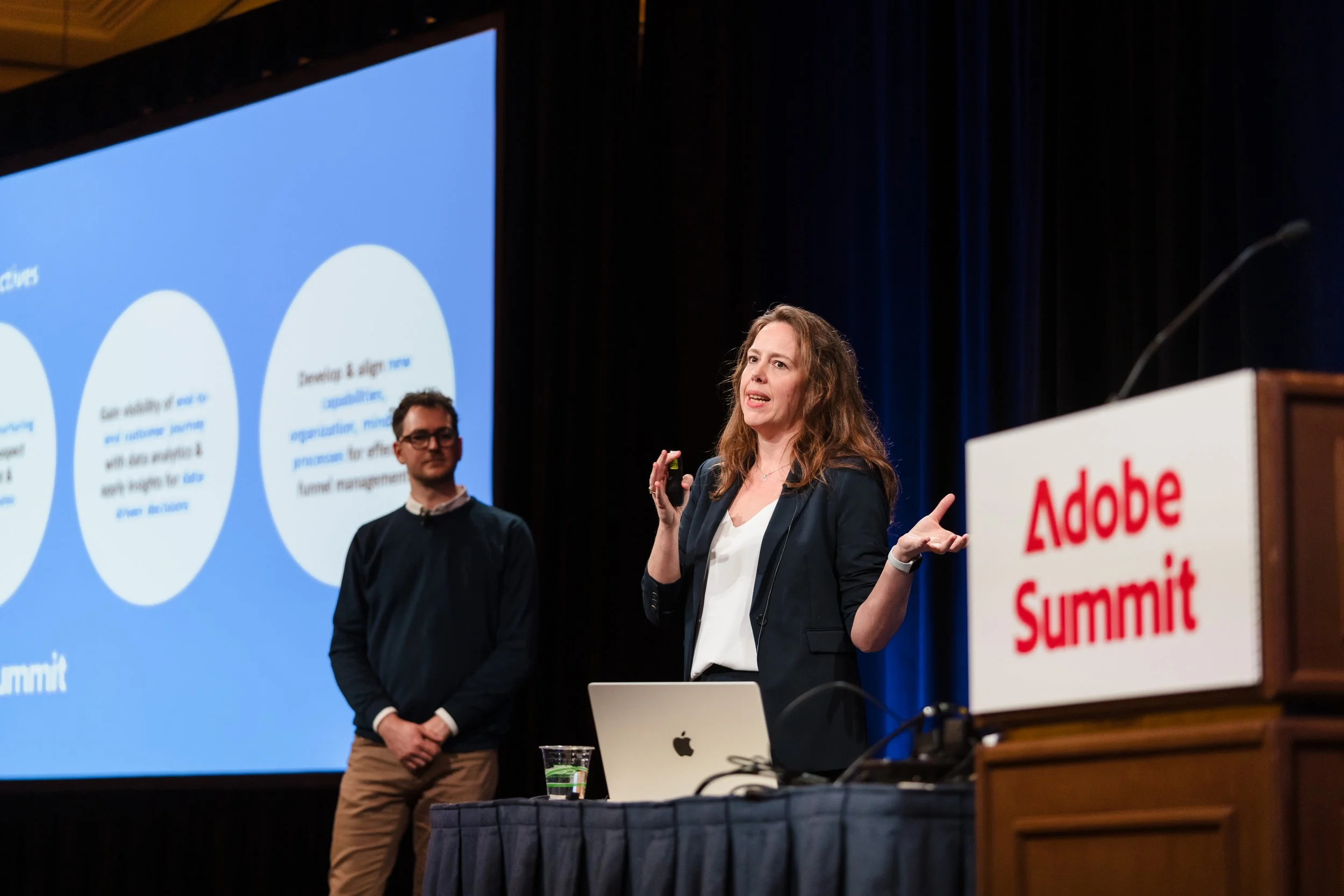 Two speakers presenting at the Adobe Summit conference. A woman with wavy brown hair is speaking at a podium with a laptop, while a man with glasses and dark hair is standing nearby listening. The background features a large screen with presentation 