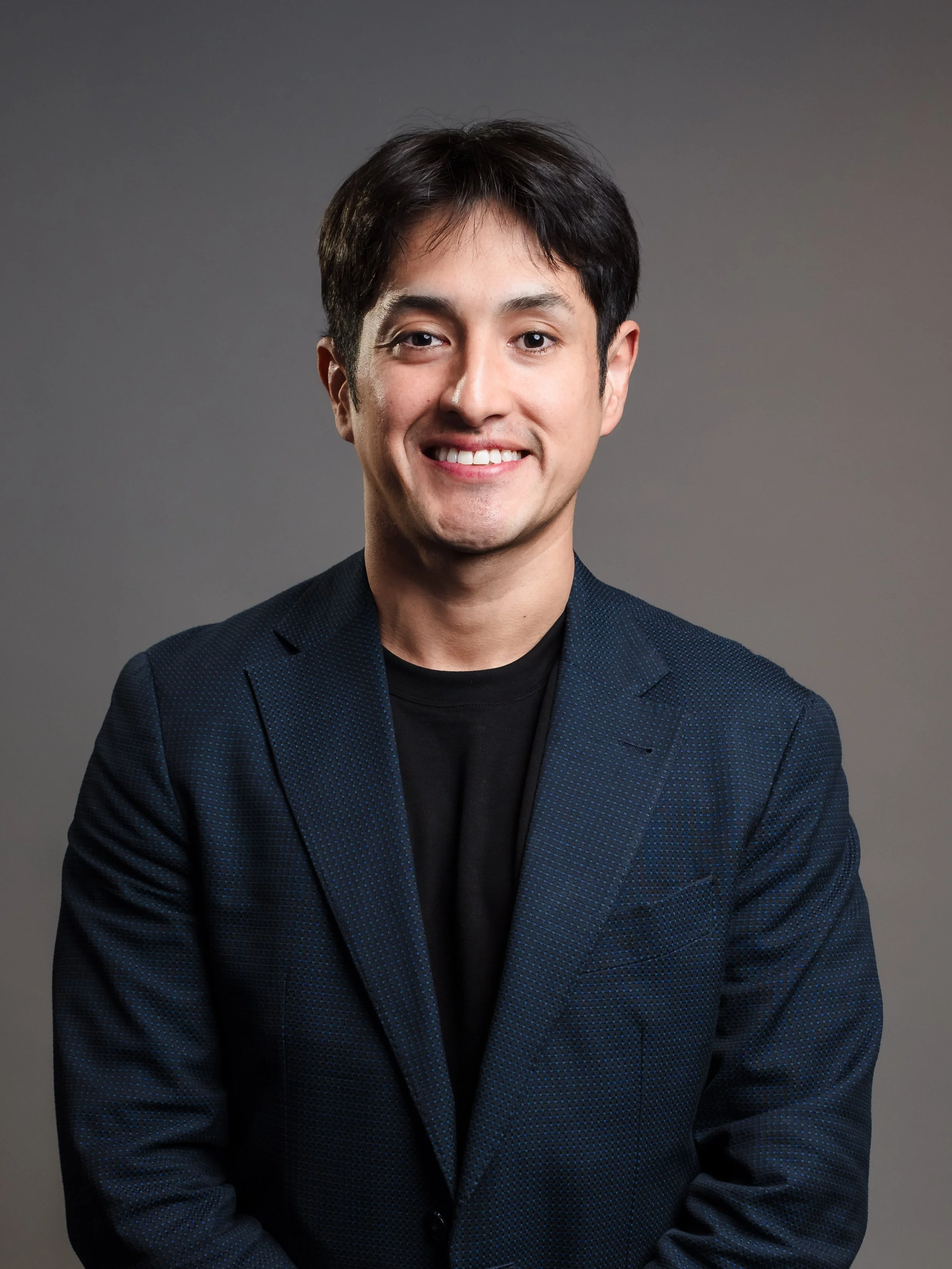 Professional portrait of a young man with dark hair, smiling, wearing a dark blazer and black shirt against a gray background.
