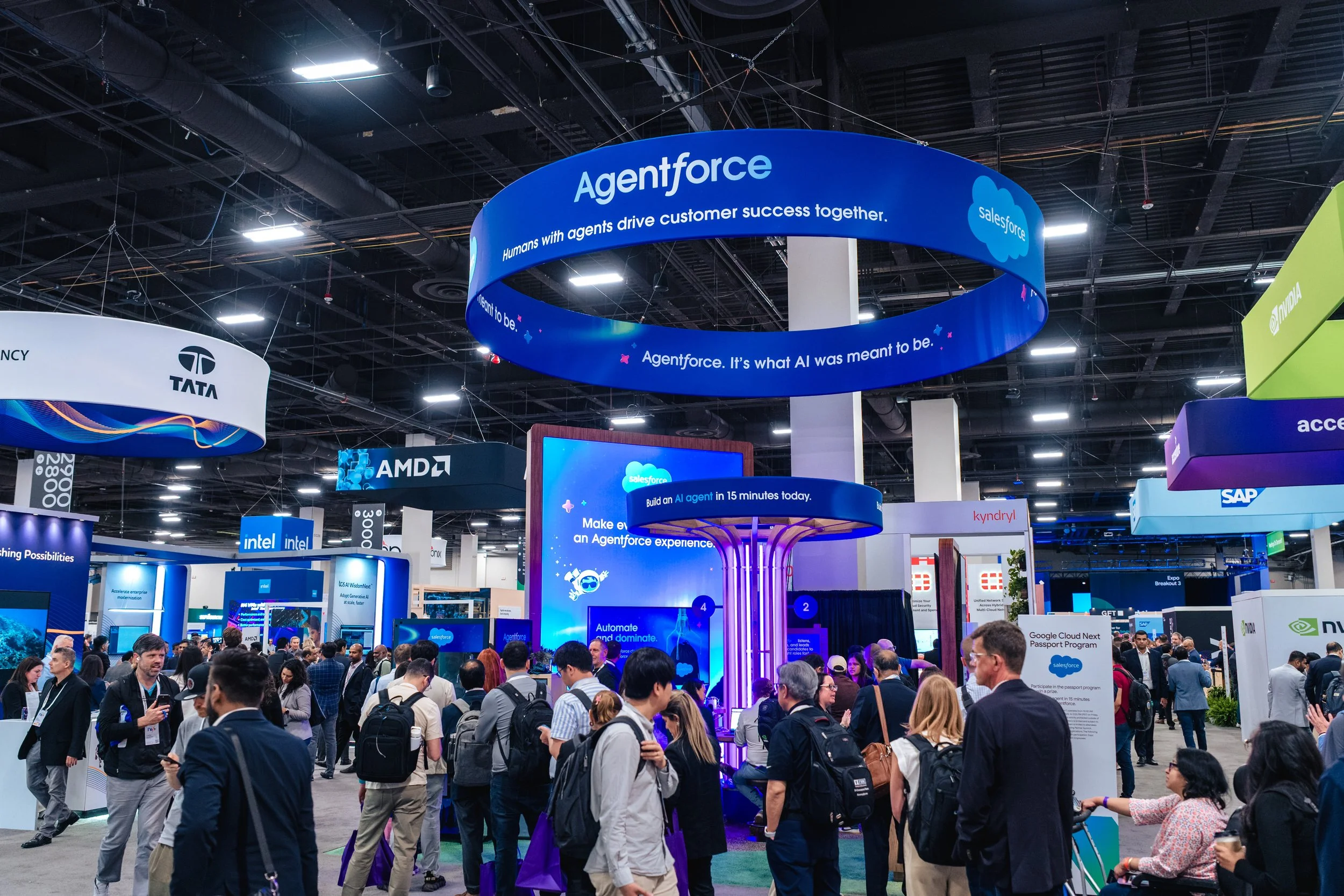 Crowd of people attending a technology expo with various booth displays, including Salesforce's Agentforce with a large circular blue sign overhead.