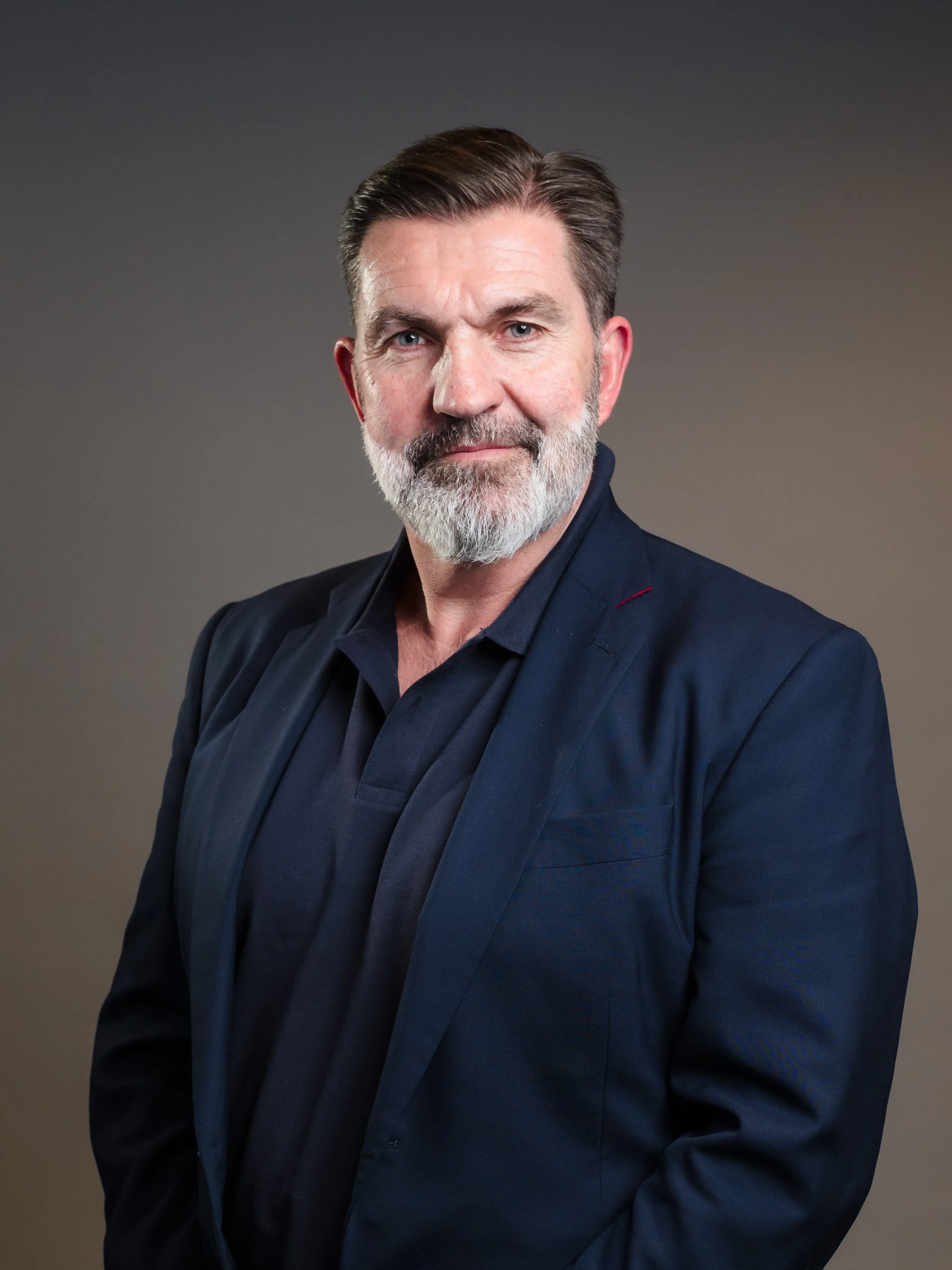 A middle-aged man with a beard and mustache, wearing a dark blazer and shirt, looking directly at the camera with a slight smile against a neutral background.