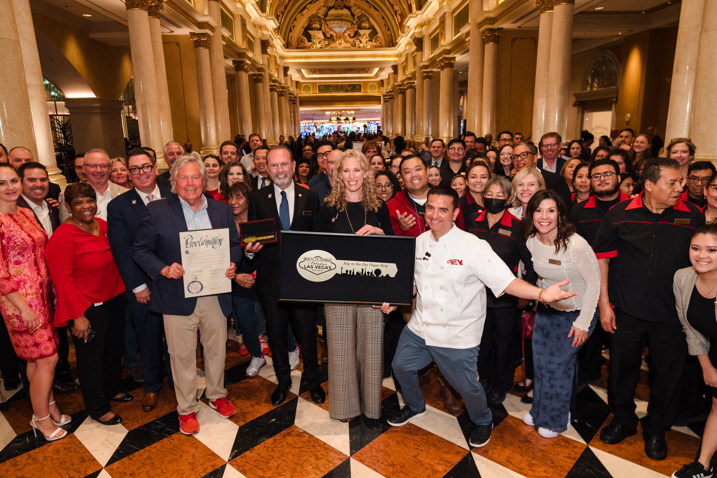A large group of people celebrating in a grand, ornate lobby, with some holding a certificate, a plaque, and a box. The group includes men and women of various ages and ethnicities, dressed in formal and casual attire, with some wearing uniforms. The