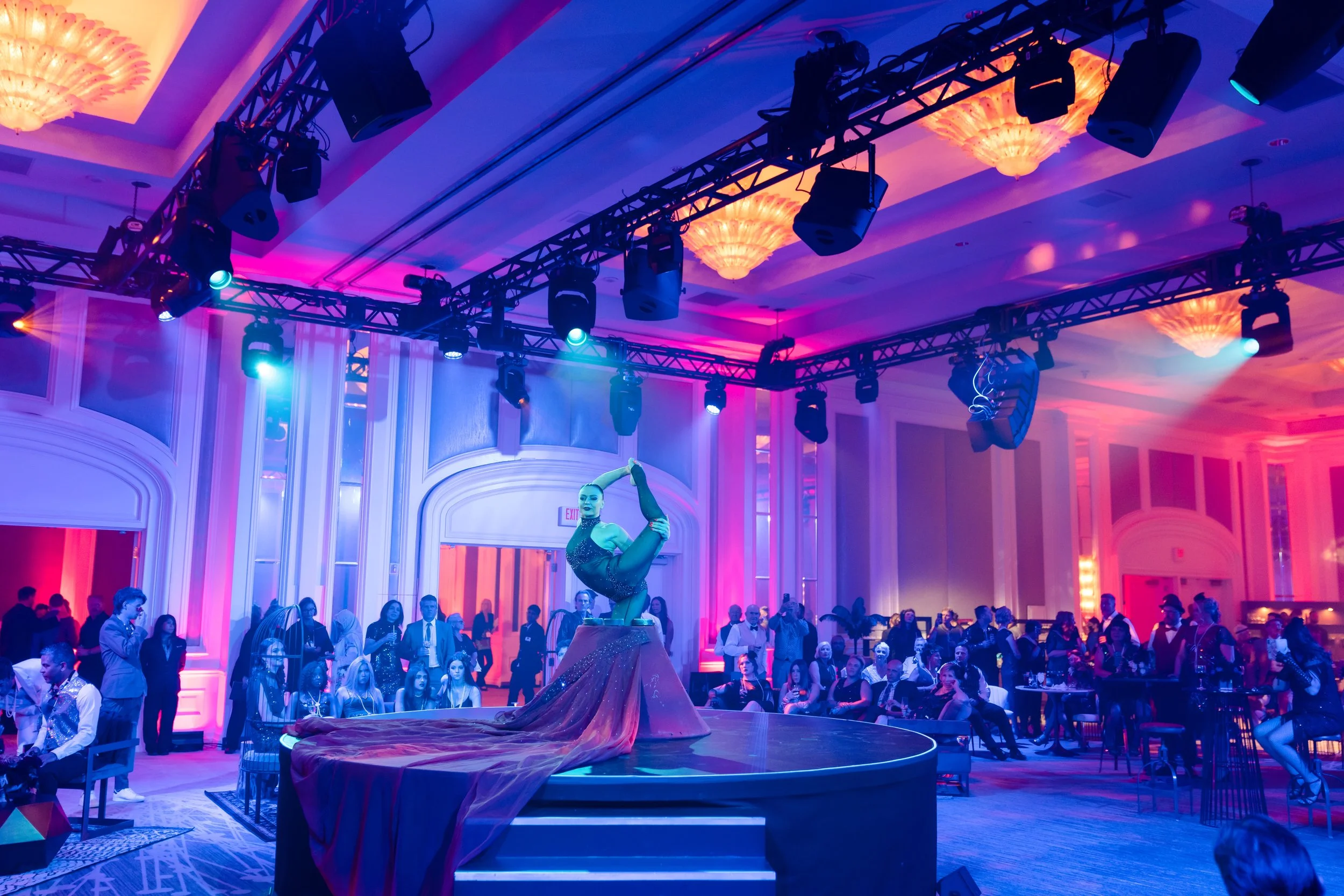 A performer in a sparkly outfit is doing a balancing act on a circular stage in a ballroom with pink and purple lighting, with an audience seated and standing around.