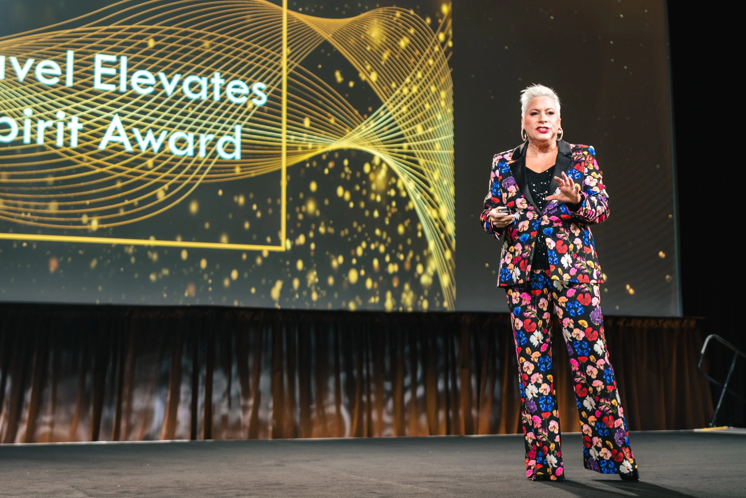 A woman with short silver hair and earrings, dressed in a colorful floral pantsuit, is speaking on stage with a large digital screen behind her displaying the words 'Level Elevates Spirit Award.'