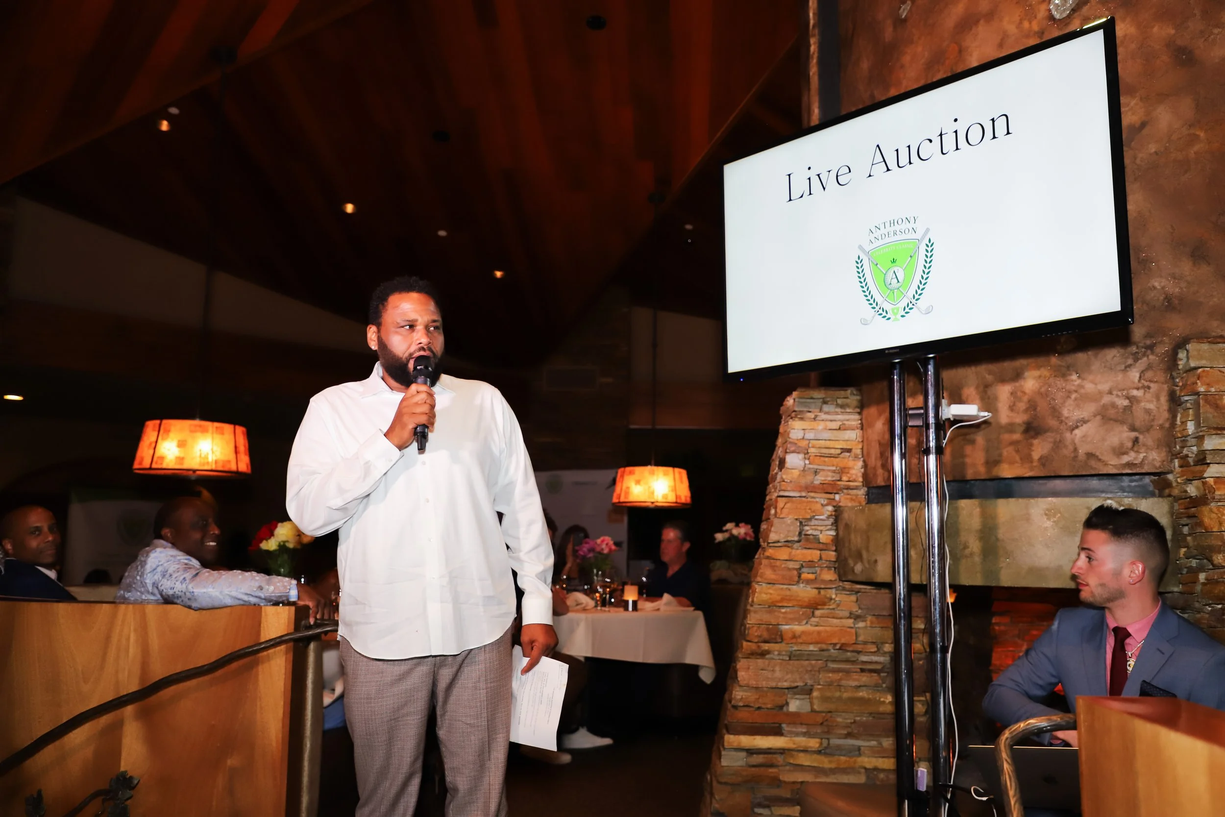 A man in a white shirt holding a microphone and speaking during a live auction in a dimly lit restaurant or event space with others seated at tables in the background.