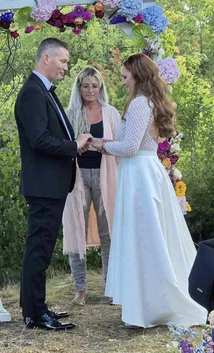 Et brudepar holder hænder under et blomsterbånd, mens en kvinde forbereder at udføre vielsen i en udendørs, grøn have.