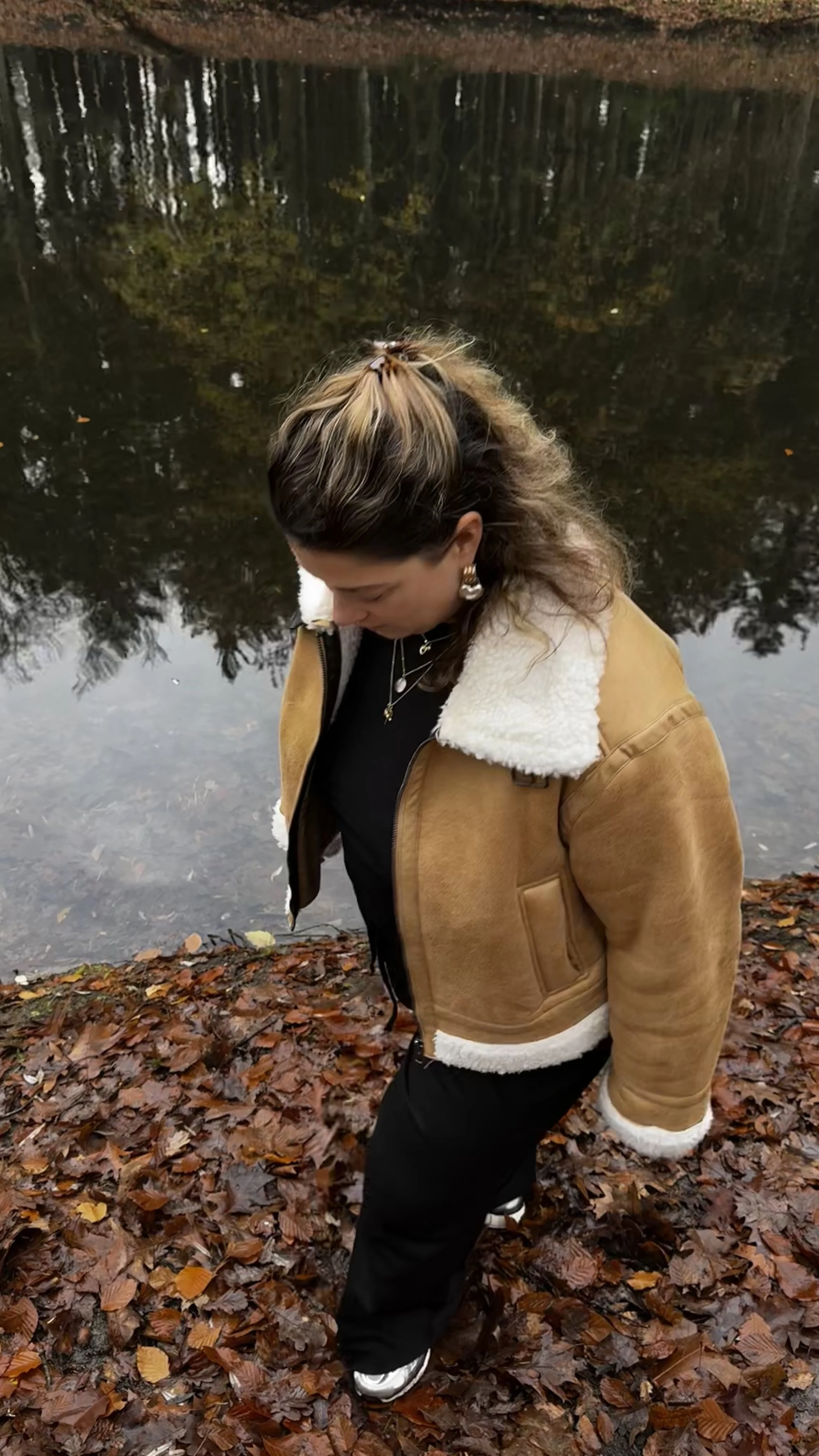 Een vrouw staat op een bedekte bladerengrond dicht bij een meer of rivier, met bomen op de achtergrond, terwijl ze naar beneden kijkt.