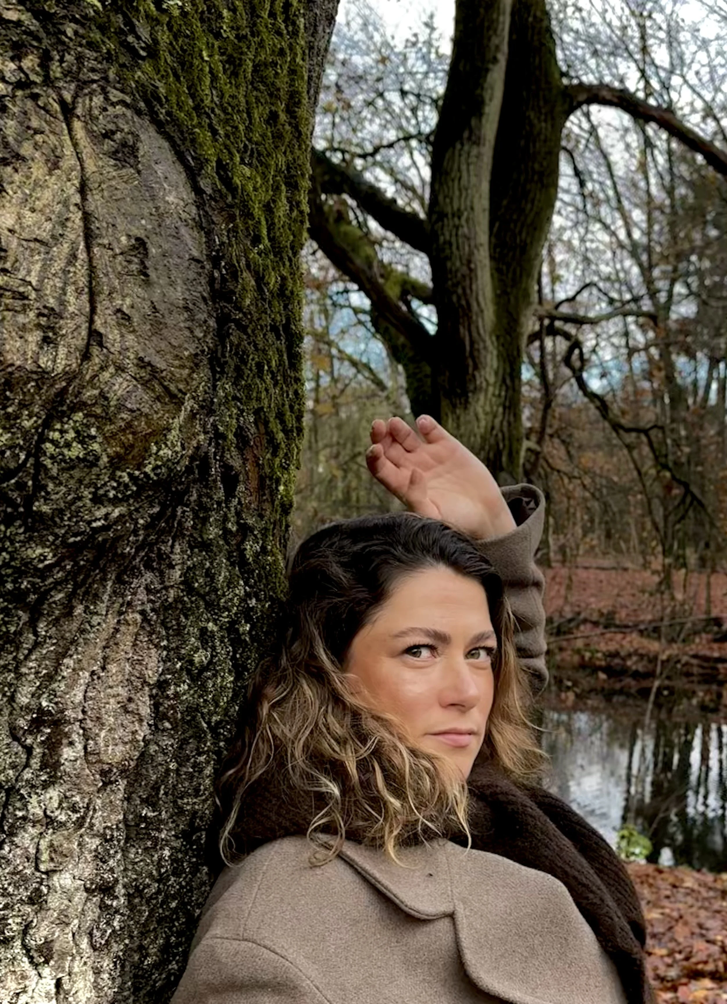 Een vrouw met golvend haar leunt tegen een boom in een bos, met een meer op de achtergrond en herfstkleuren in de omgeving.