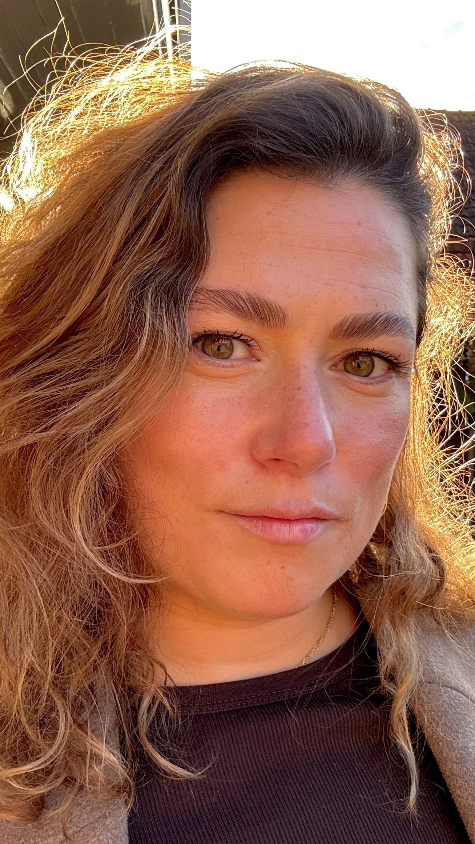 Close-up van een vrouw met golvende bruin haar en groene ogen, die in de zon kijkt.