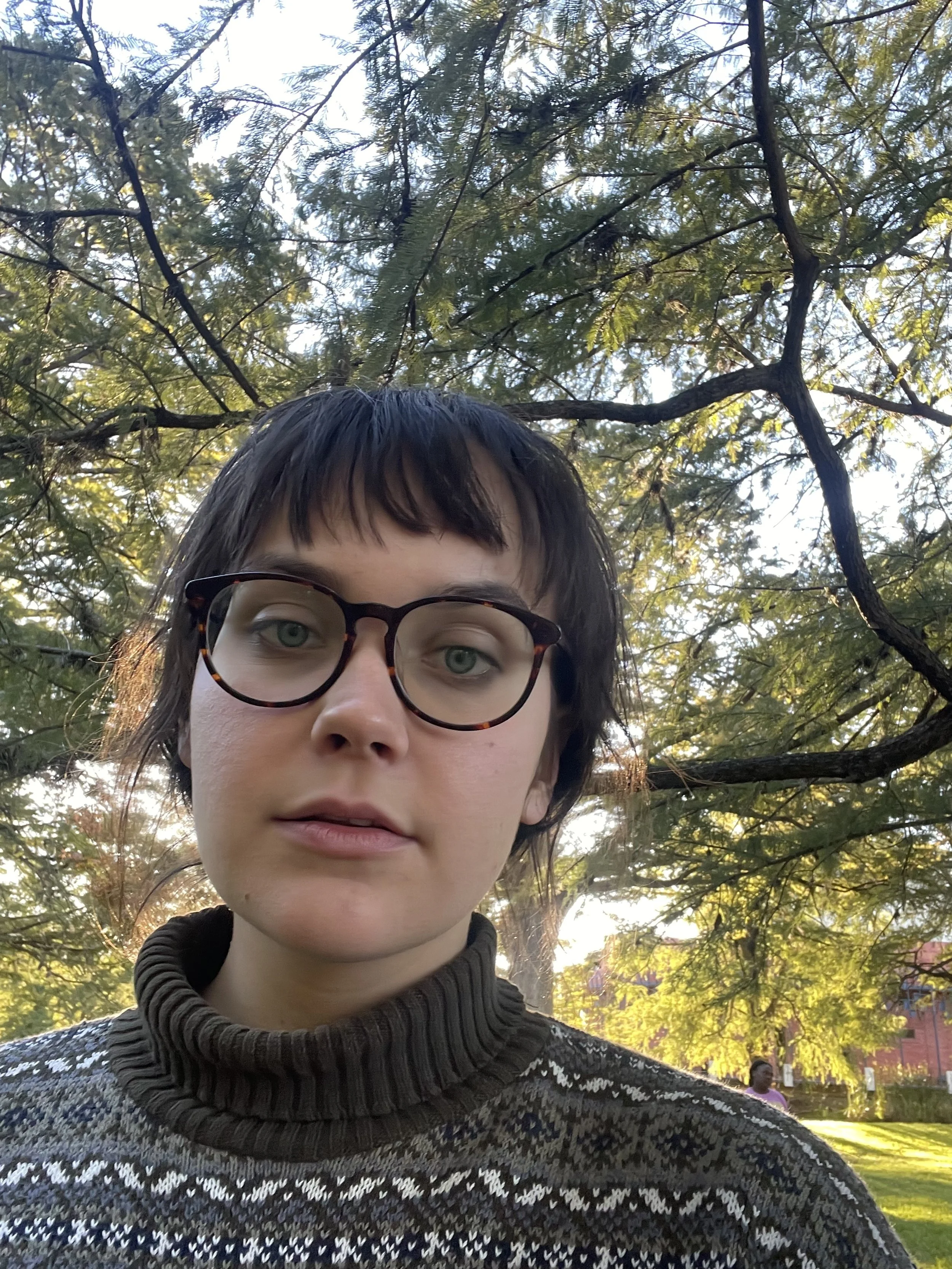 A selfie of Devon wearing glasses and sweater in front of trees