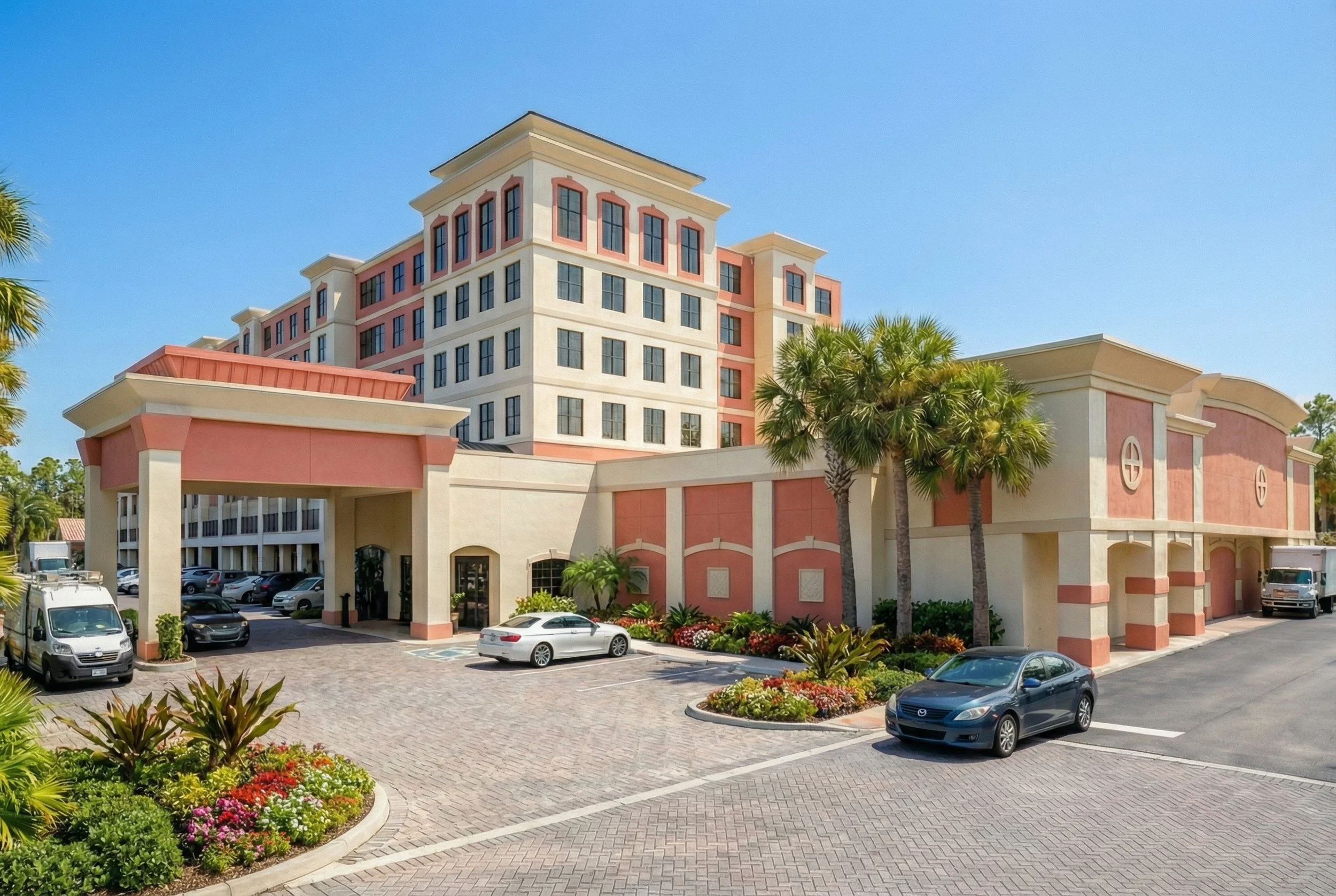 A multi-story building with pink and cream colors, surrounded by palm trees and a parking lot with several cars, under a clear blue sky.