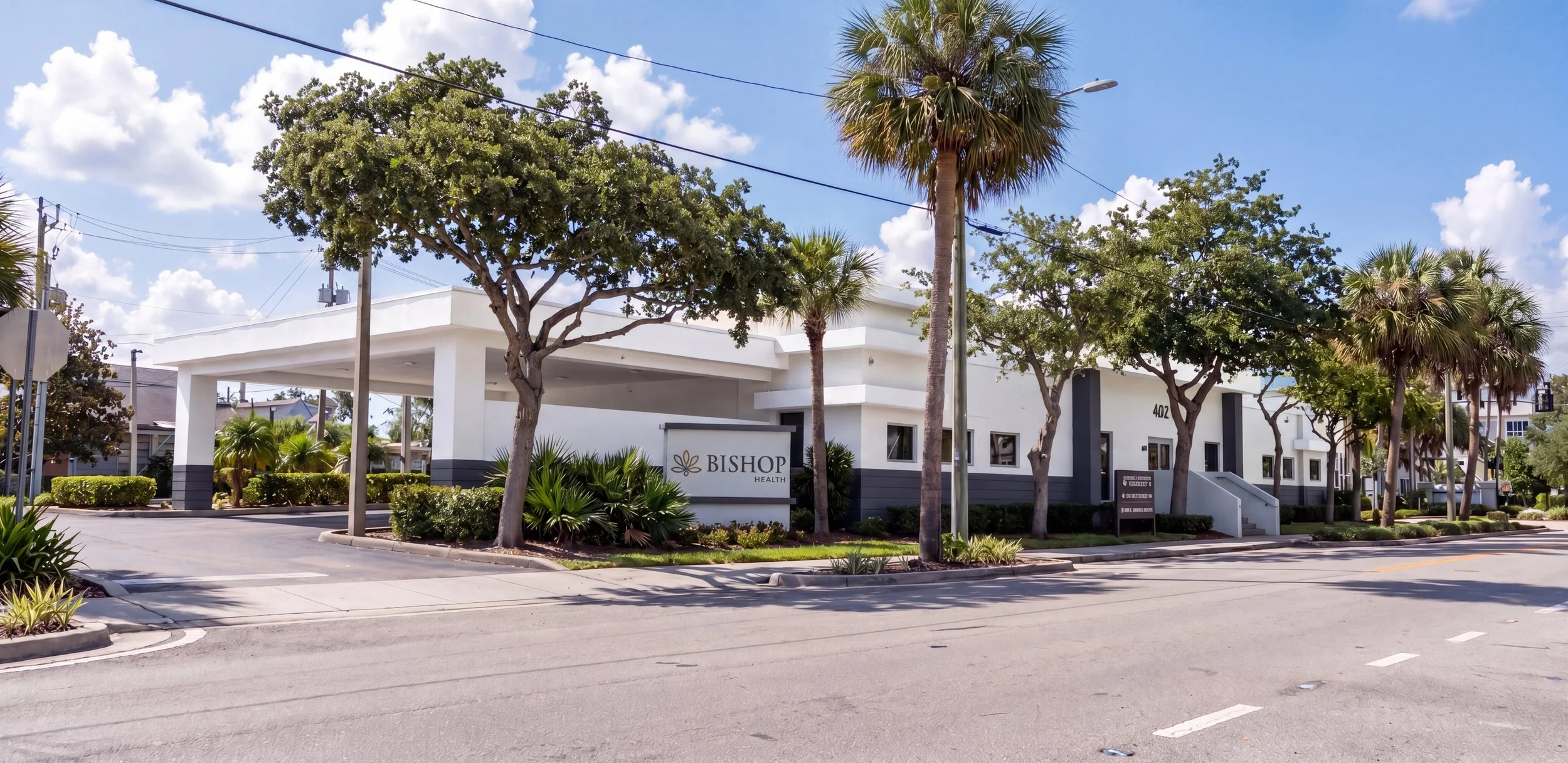 A modern white building with a flat roof and the sign 'BISHOP HEALTH' in front, surrounded by palm trees and green shrubs under a blue sky with white clouds.