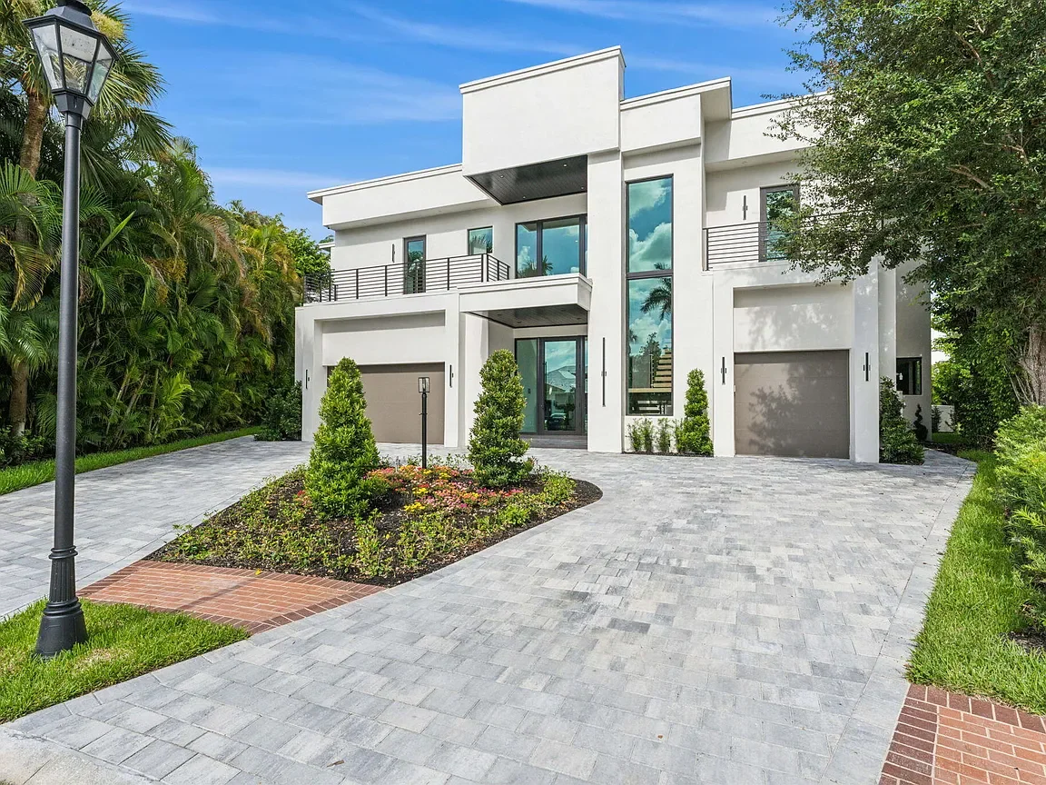 Modern white house with large glass windows and driveway, surrounded by greenery and landscaped garden.